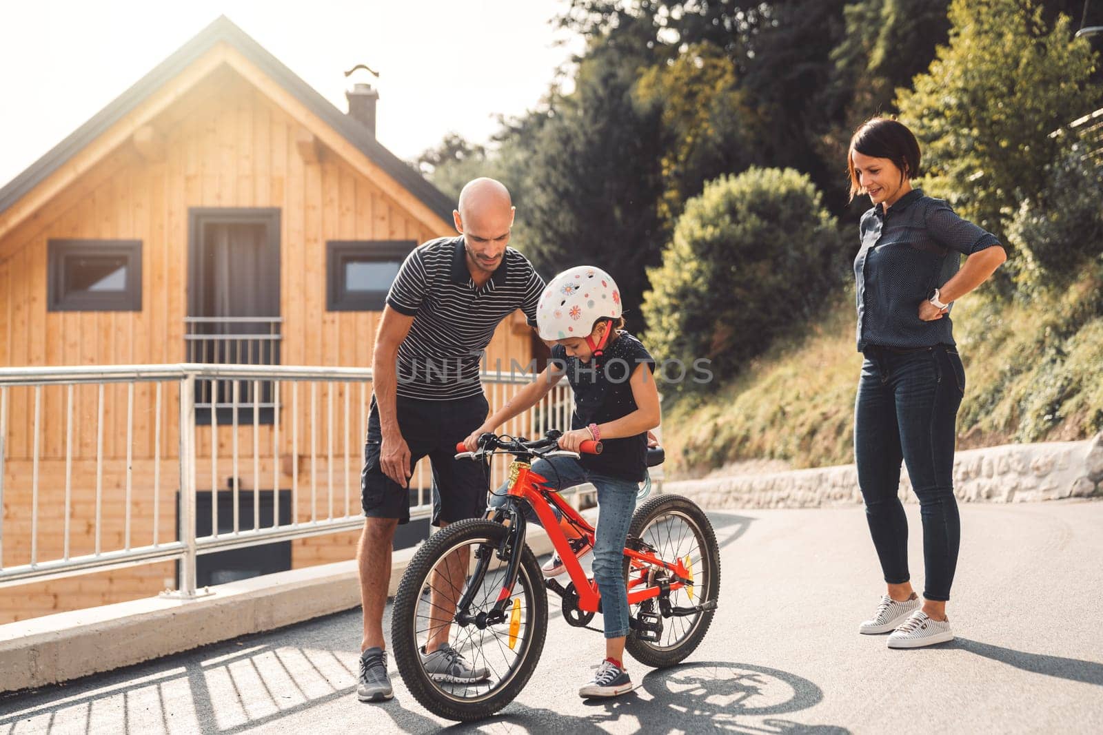 Dad teaching her young daughter how to ride a bike, while mom stands by looking at them by VisualProductions