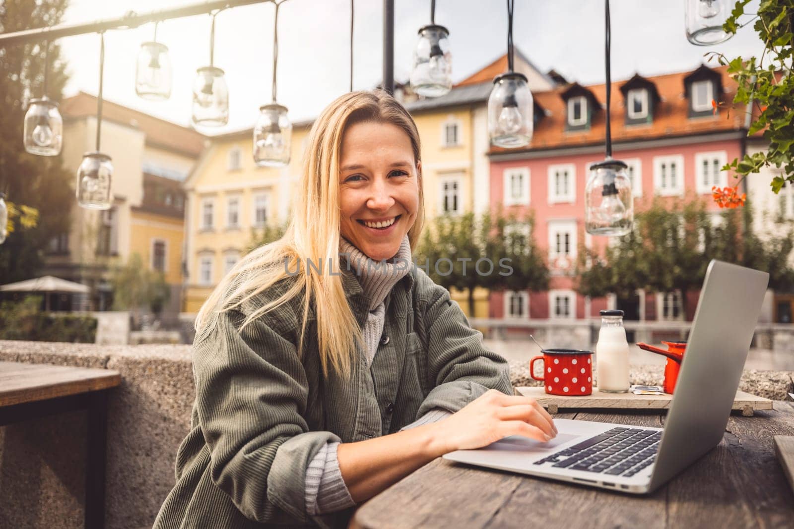 Cheerful blonde woman sitting at a cafe outside, working on her laptop, smiling at the camera by VisualProductions