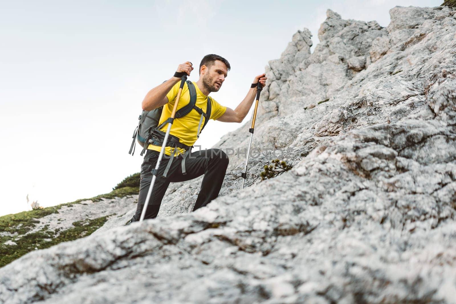 Caucasian mountaineer in a yellow t-shirt walking up the rocky mountain with hiking poles, low angle view by VisualProductions