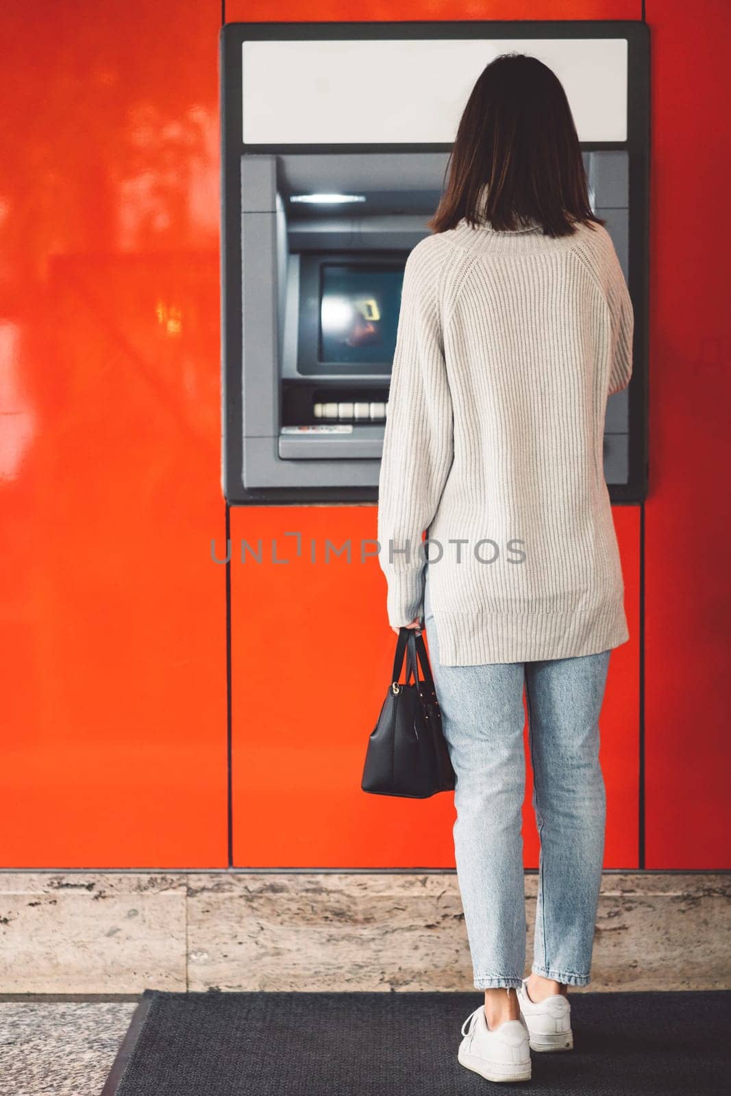 Vertical photo, back view of a woman standing in front of an ATM on a red wall, making a cash withdrawal by VisualProductions