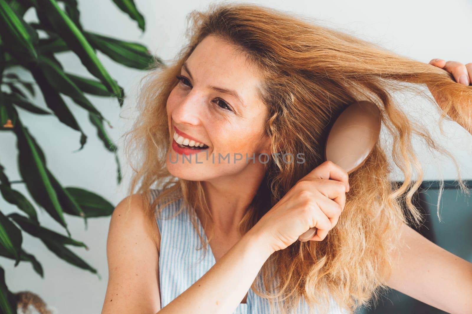 Waist up portrait smiling woman brushing her blonde curly hair with a hair brush by VisualProductions