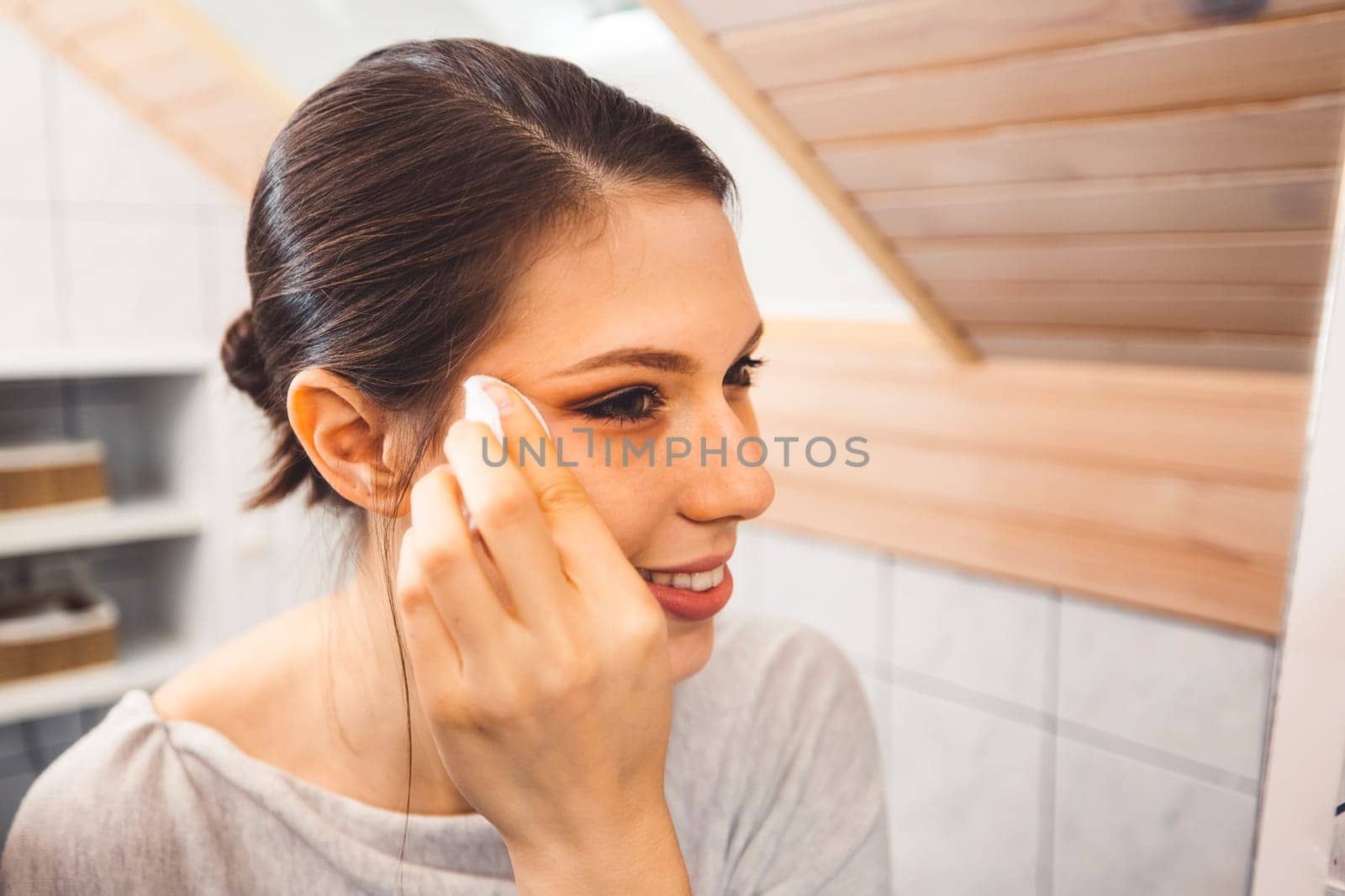 Smiling cheerful young woman cleaning make up off her face late at night in her bathroom by VisualProductions
