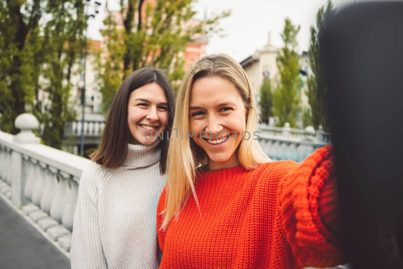 Couple of best friends in colorful sweaters taking a selfie on a bridge in the city, trees in the background by VisualProductions