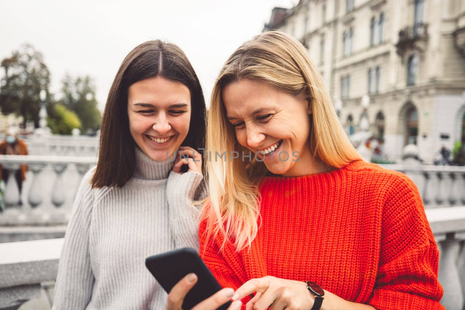 Waist up portrait two smiling young women in red and beige sweater walking around the city looking down at their phone by VisualProductions