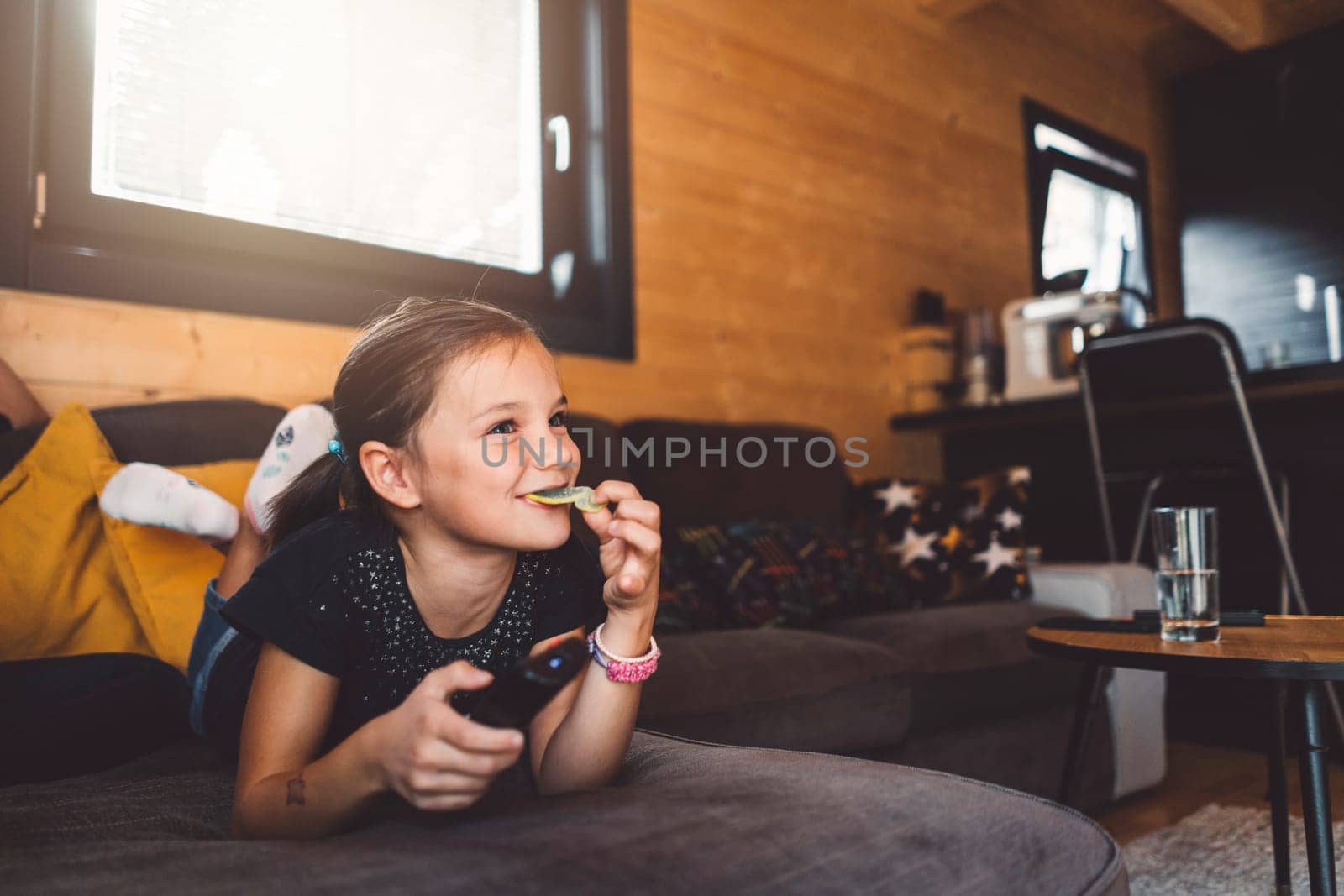 Cheerful little girl having snacks while watching her favorite show on the television by VisualProductions