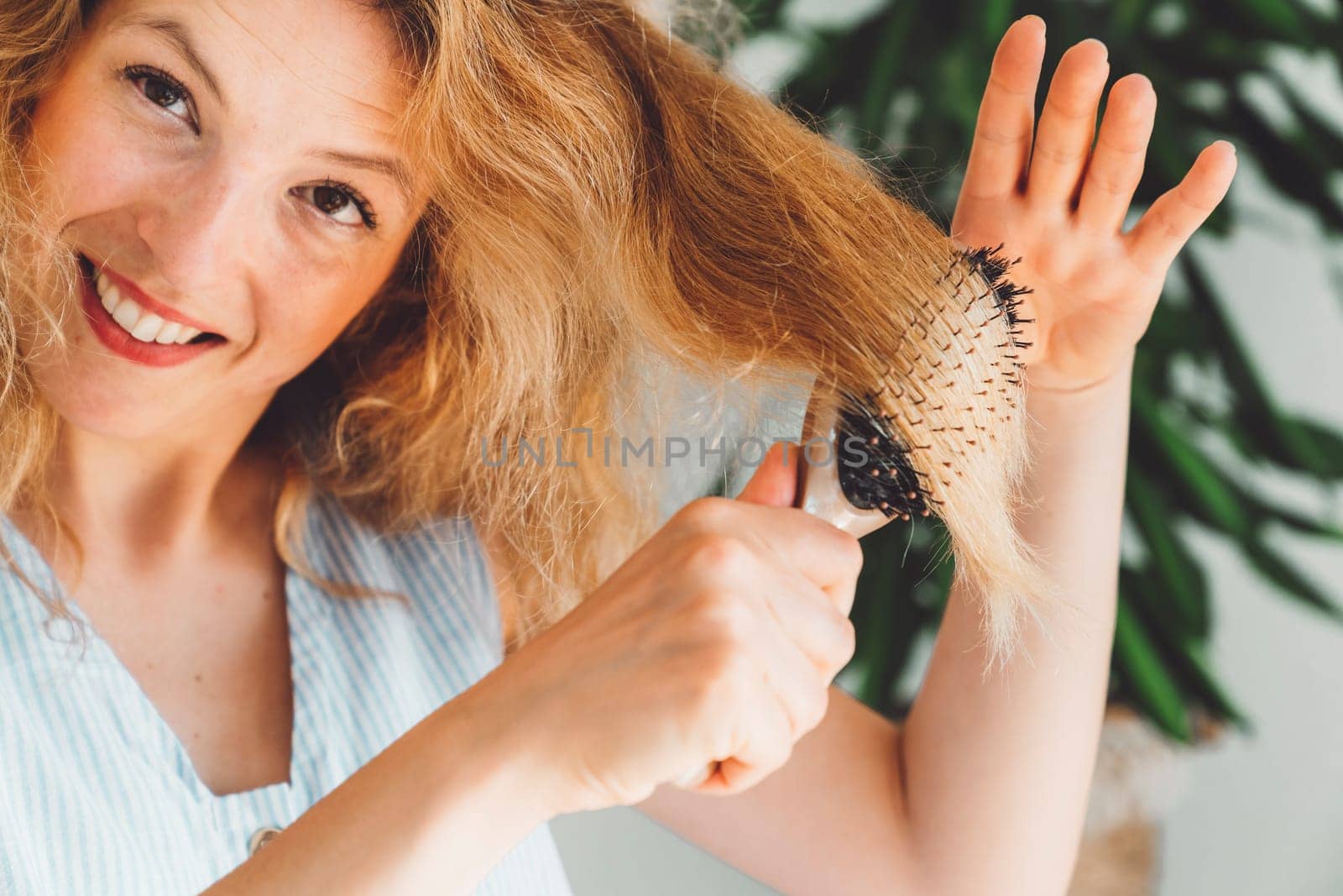 Smiling woman brushing her blonde hair with a brush, doing her hairstyle by herself at home by VisualProductions
