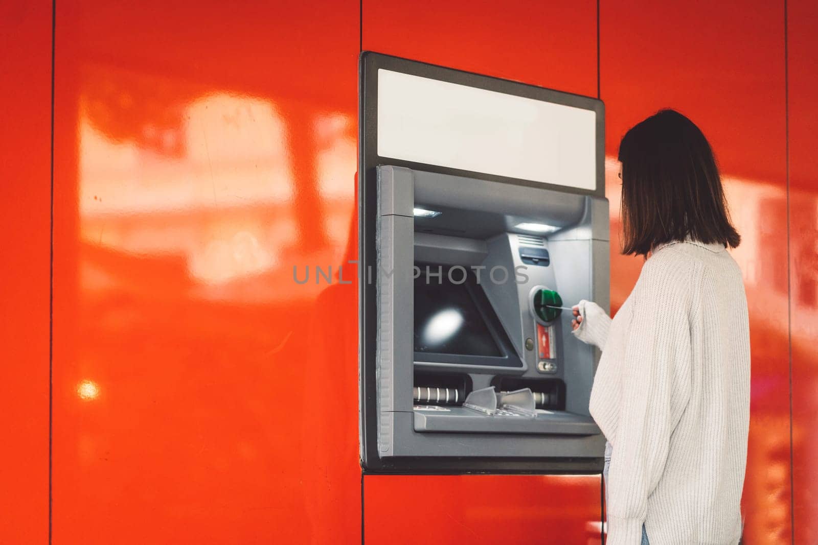 Side view of a woman inserting a card in the ATM on a red wall by VisualProductions