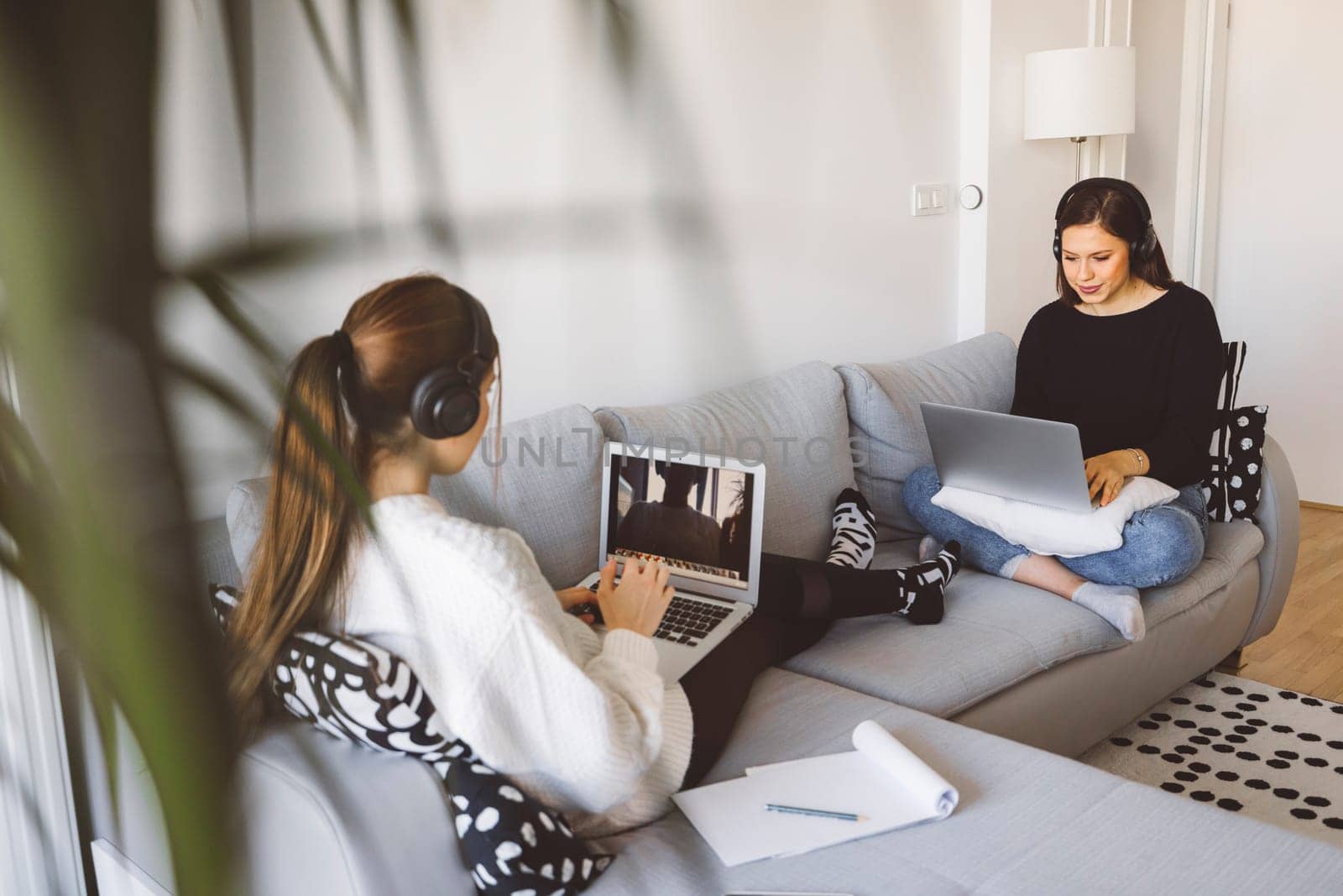 Two young women students studying on their laptops on the couch in their apartment each with their headphones on by VisualProductions