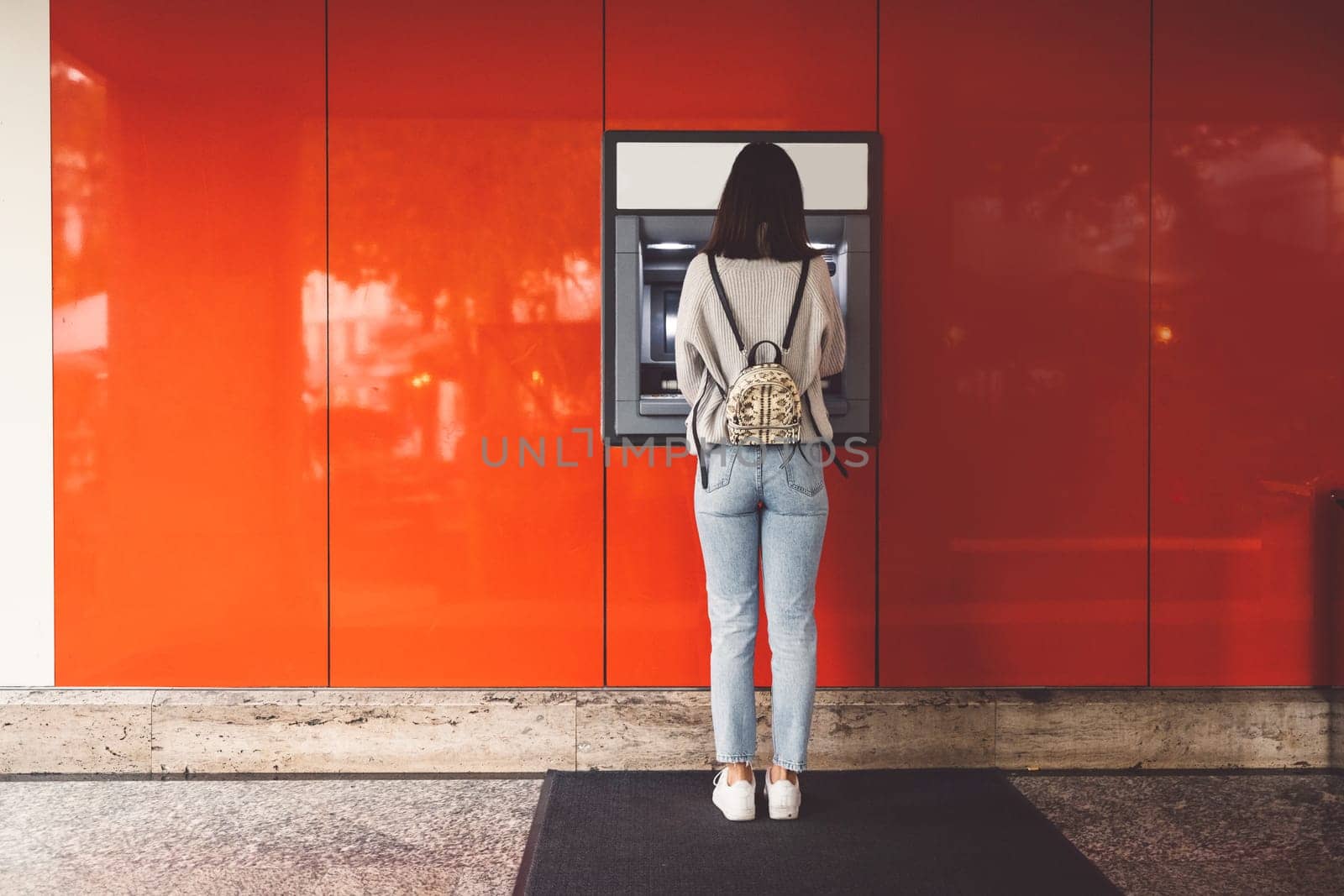 Back view of young woman standing in front of an ATM machine on a red wall by VisualProductions