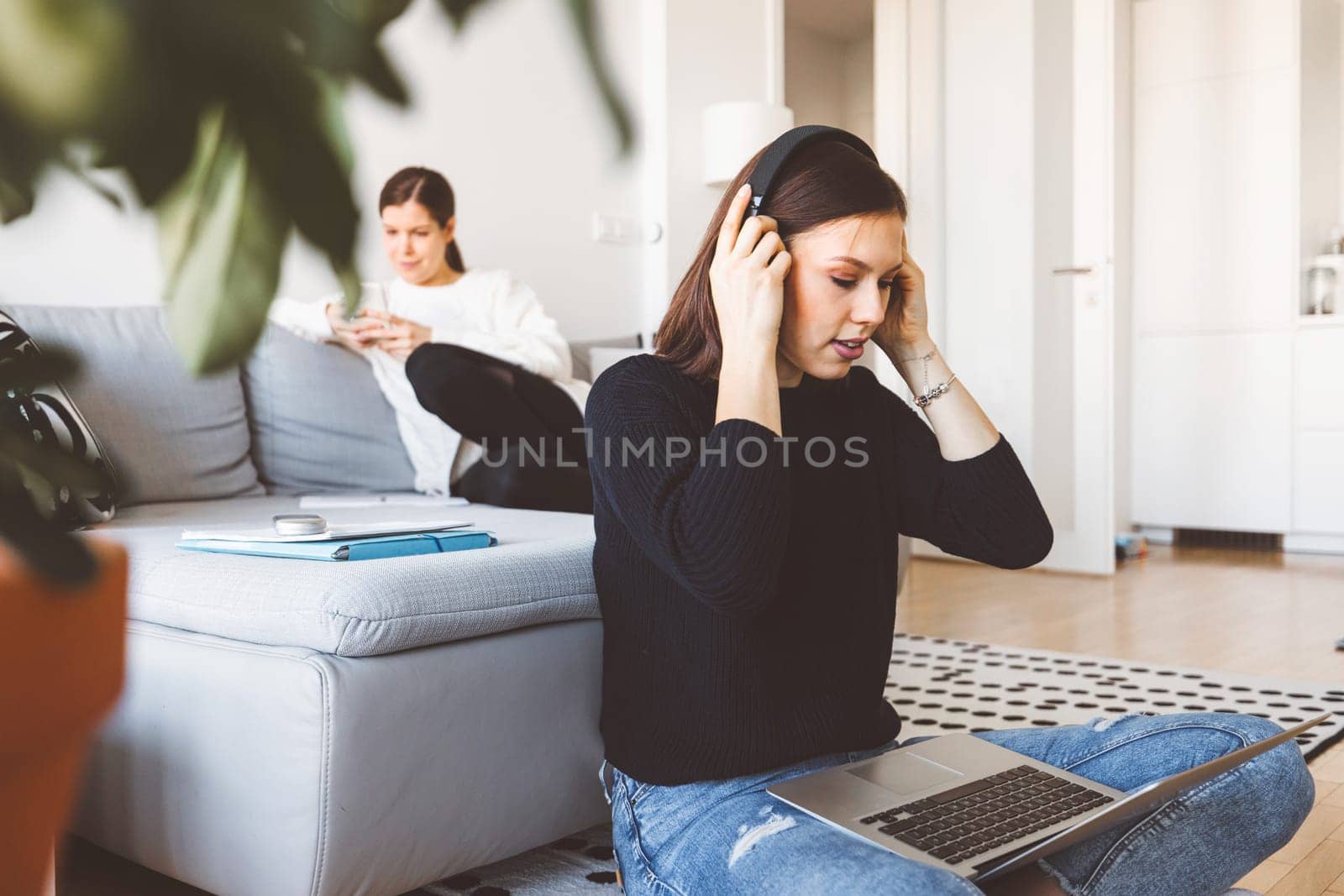 Two young women college students in their apartment studying on laptops, one in front putting head phones on by VisualProductions