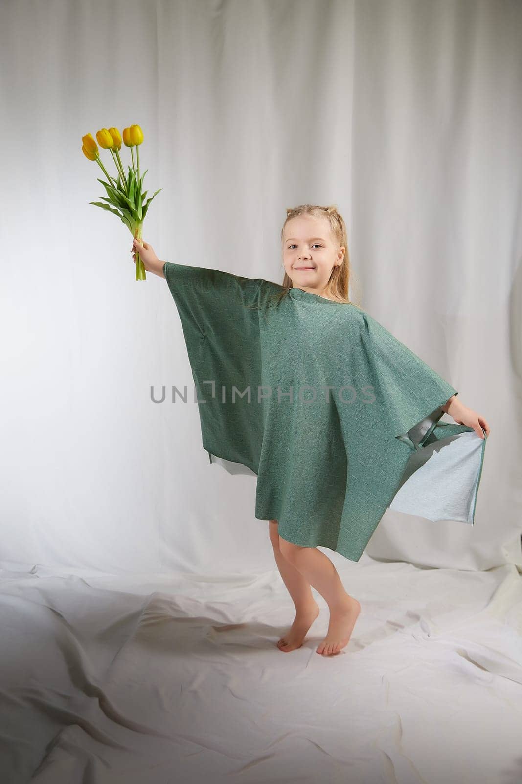 Portrait of a little blonde girl with bouquet of spring yellow flowers on a light background. Child in green dress holding a bouquet of tulips in hands. Spring concept