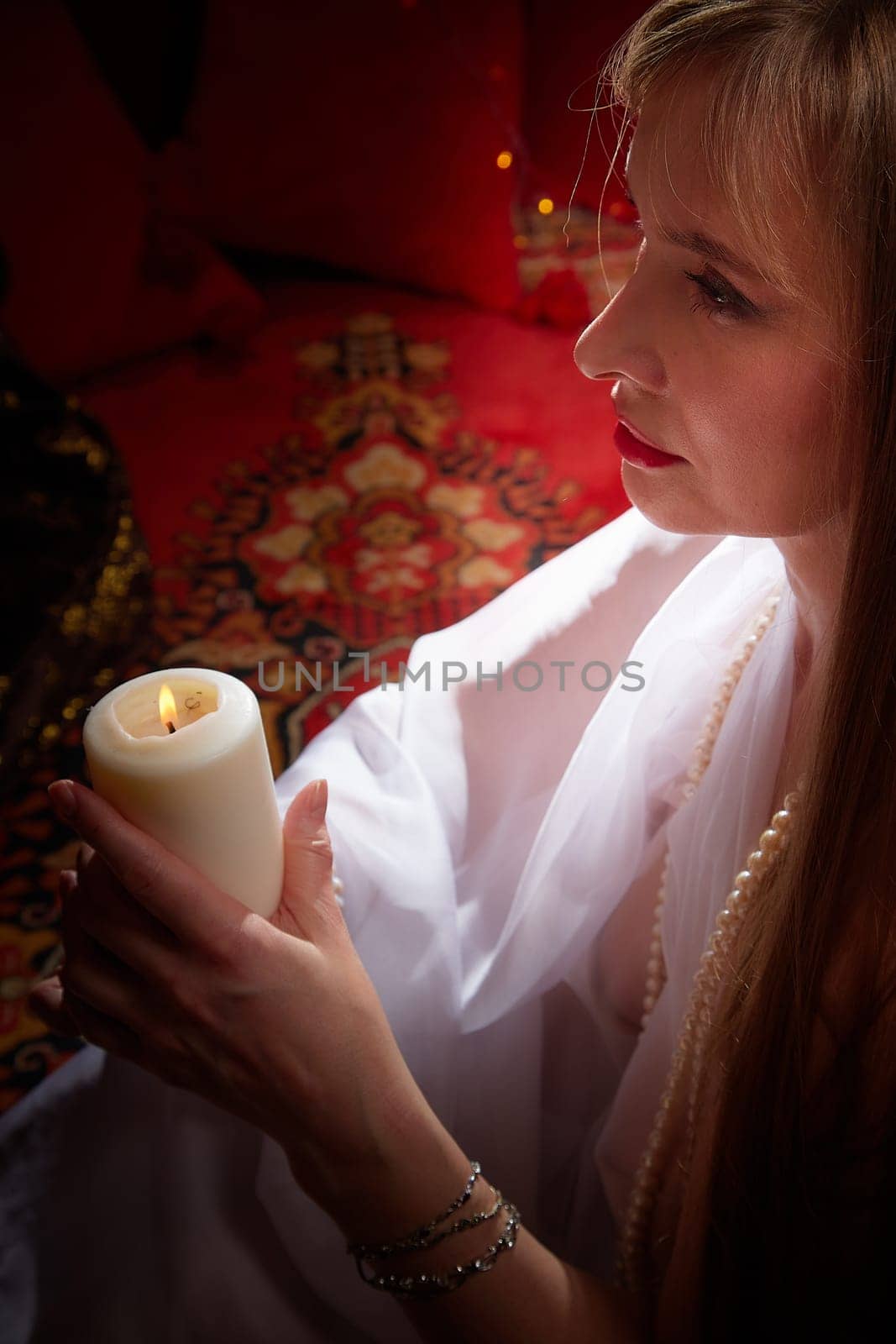 Beautiful arabian girl with candles in red room full of rich fabrics and carpets in sultan harem. Photo shoot of woman an oriental style odalisque. Model poses in sari as caring wife and hostess