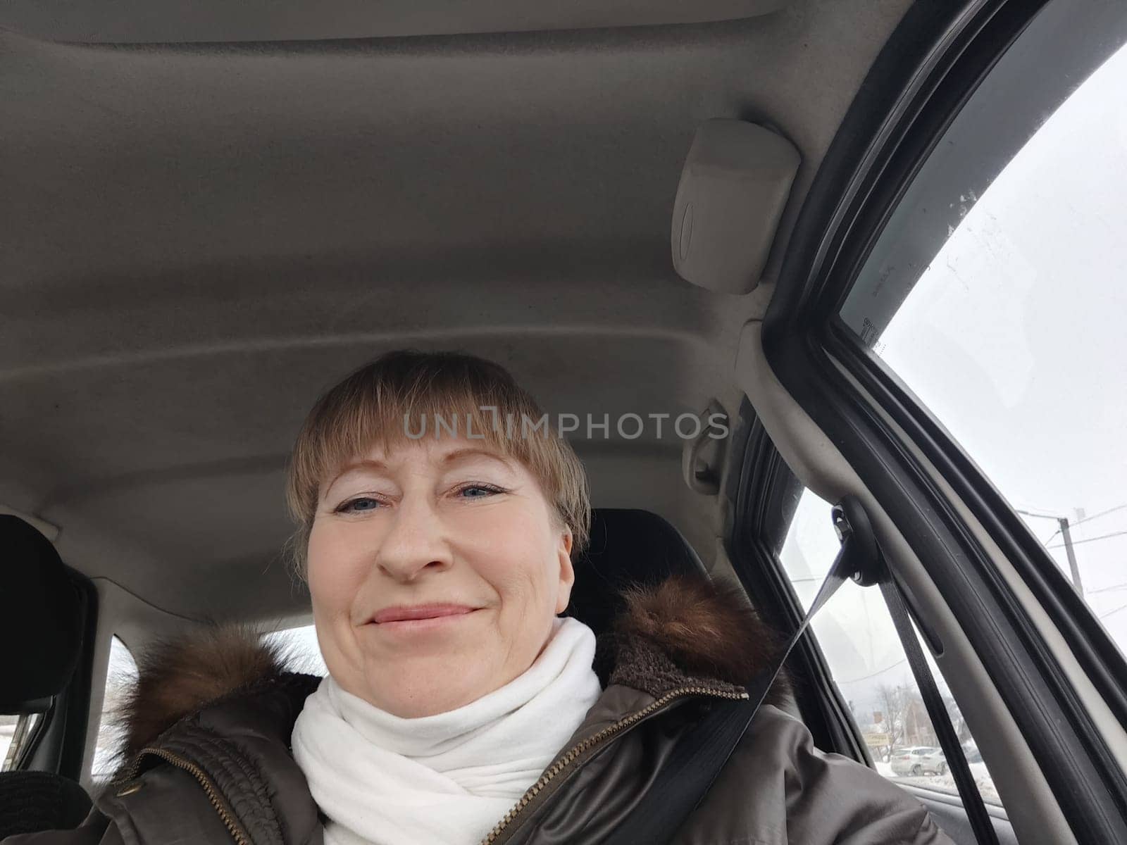 Portrait of adult cute funny blonde woman in warm clothes taking a selfie inside the car and driving it. The concept of a pleasant trip in spring, autumn, winter by keleny