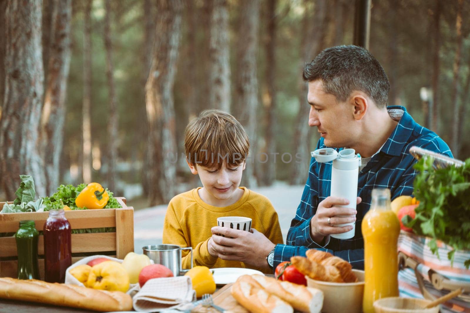 Father dad and school kid boy child having a picnic in the forest camping site with vegetables, juice, coffee, and croissants. Wooden crate with fresh organic veggies surrounded with bread baguettes. by Ostanina