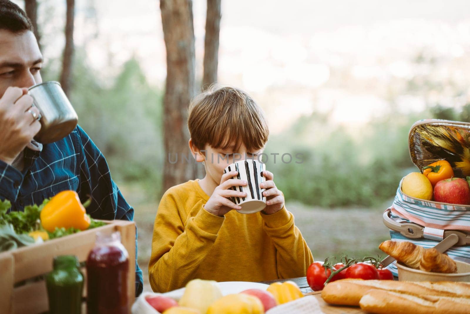 Father dad and school kid boy child having a picnic in the forest camping site with vegetables, juice, coffee, and croissants. Wooden crate with fresh organic veggies surrounded with bread baguettes. by Ostanina