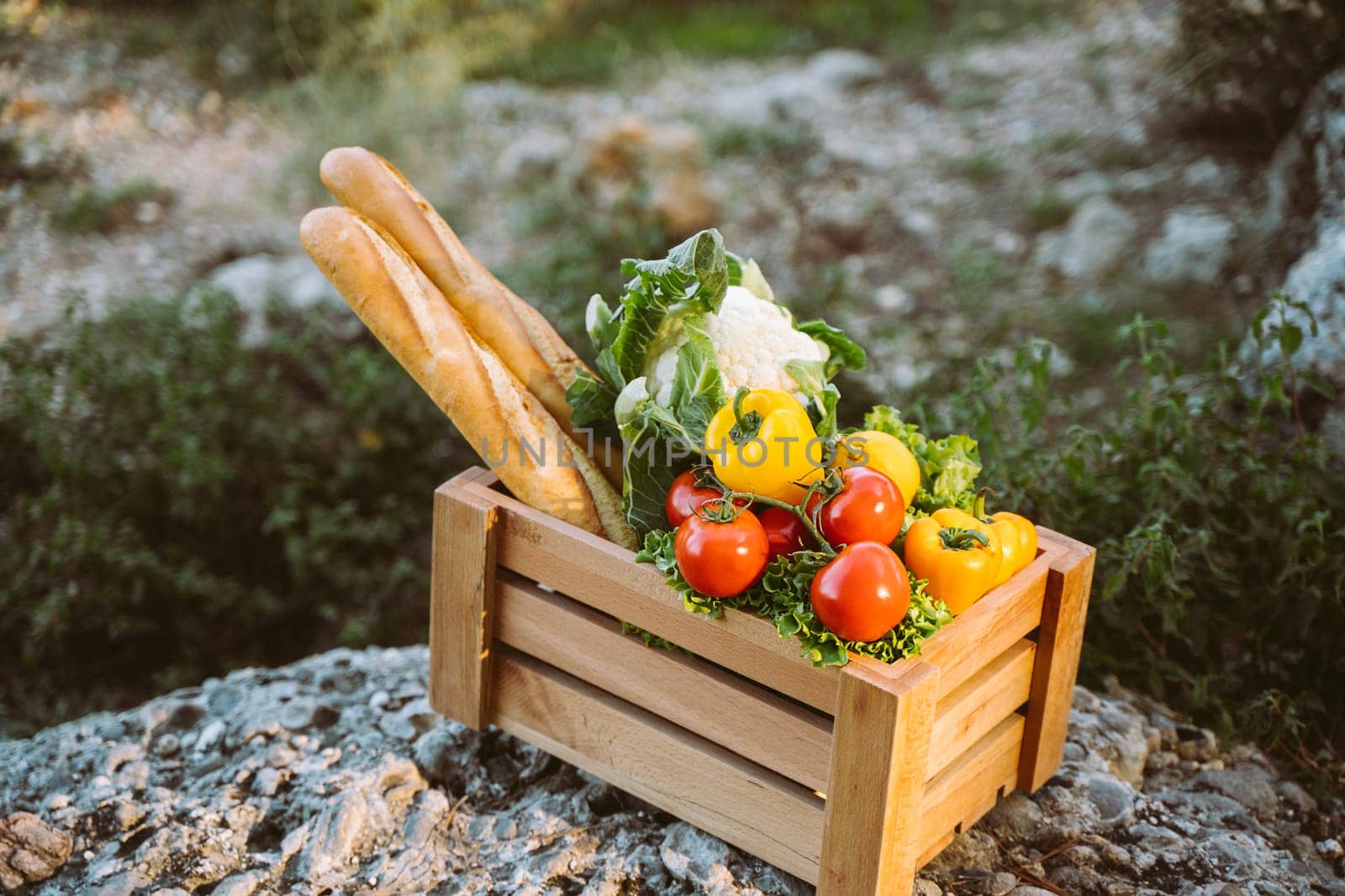 Wooden crate with farm market organic vegetables and baguettes standing on a stone. Closeup photo of frame crate with vegan food, tomatoes, bread, cauliflower, and sweet pepper