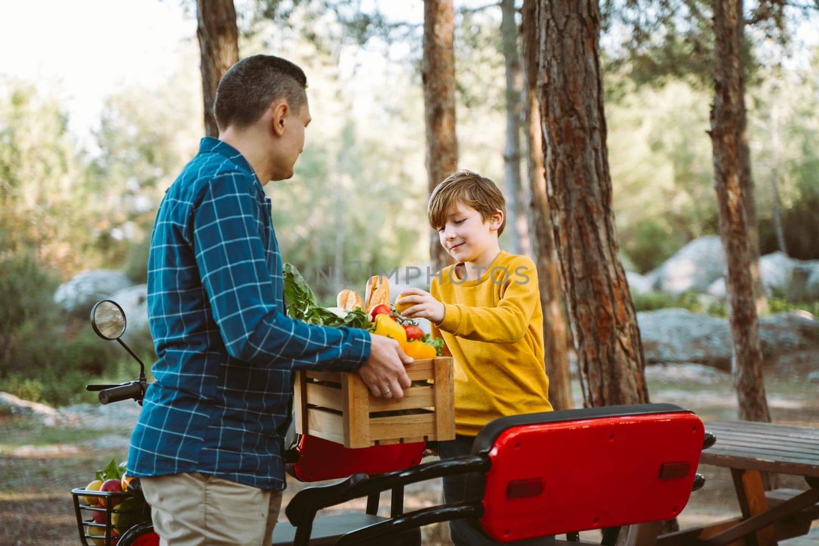 Farmer father and school boy kid delivered wooden crates with vegetables to camp site for picnic. Dad man male and son tourists unloading groceries from electric tricycle vehicle in the forrest