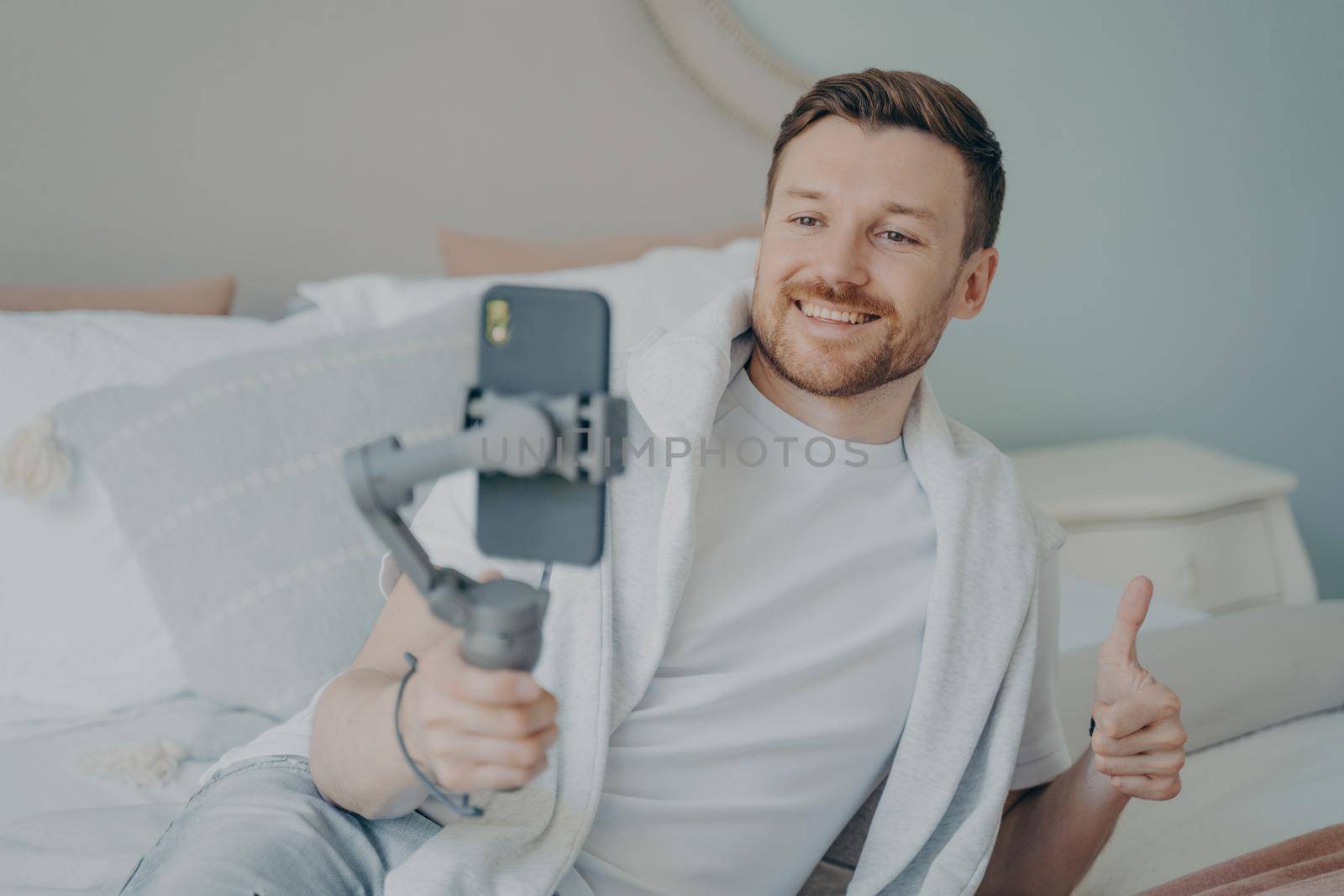 Young positive bearded man showing thumb up while doing online call on smartphone at home by vkstock