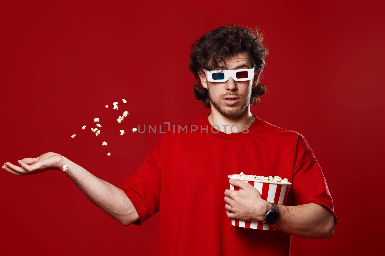 young surprised man in 3d glasses, red t-shirt throwing up popcorn on red background