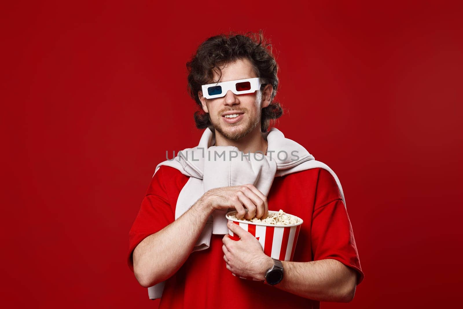 young bearded man in 3d-glasses eating popcorn on red background