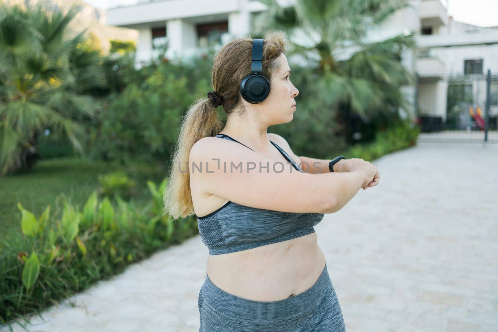 Adorable fat woman in tracksuit is engaged in fitness outdoor side view portrait. Young overweight woman lunges outdoors on warm summer day. Healthy lifestyle and weight loss.