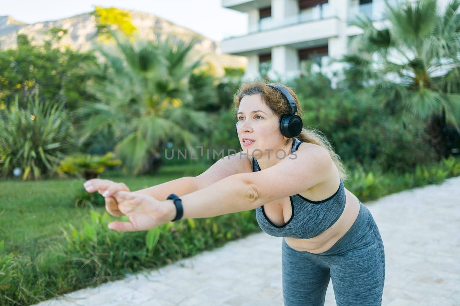 Adorable fat woman in tracksuit is engaged in fitness outdoor side view portrait. Young overweight woman lunges outdoors on warm summer day. Healthy lifestyle and weight loss.