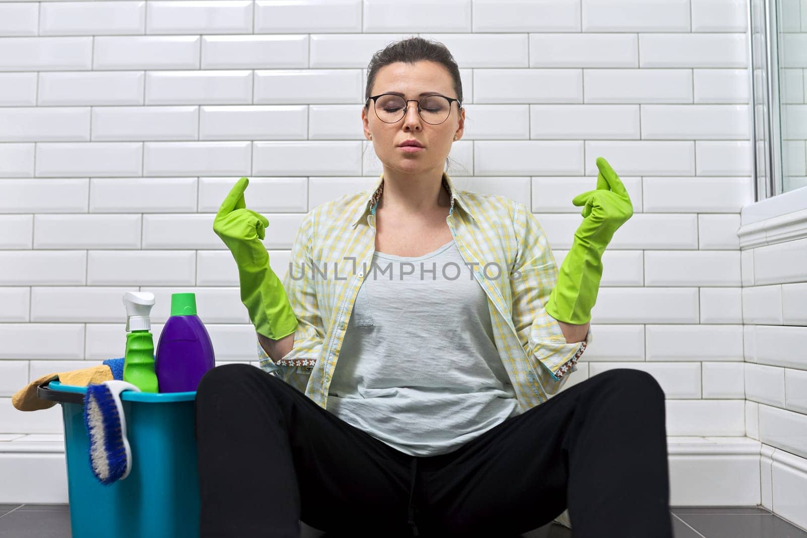 Tired woman sitting in bathroom on floor and meditating. Female in clean room with bucket of detergent resting in meditation