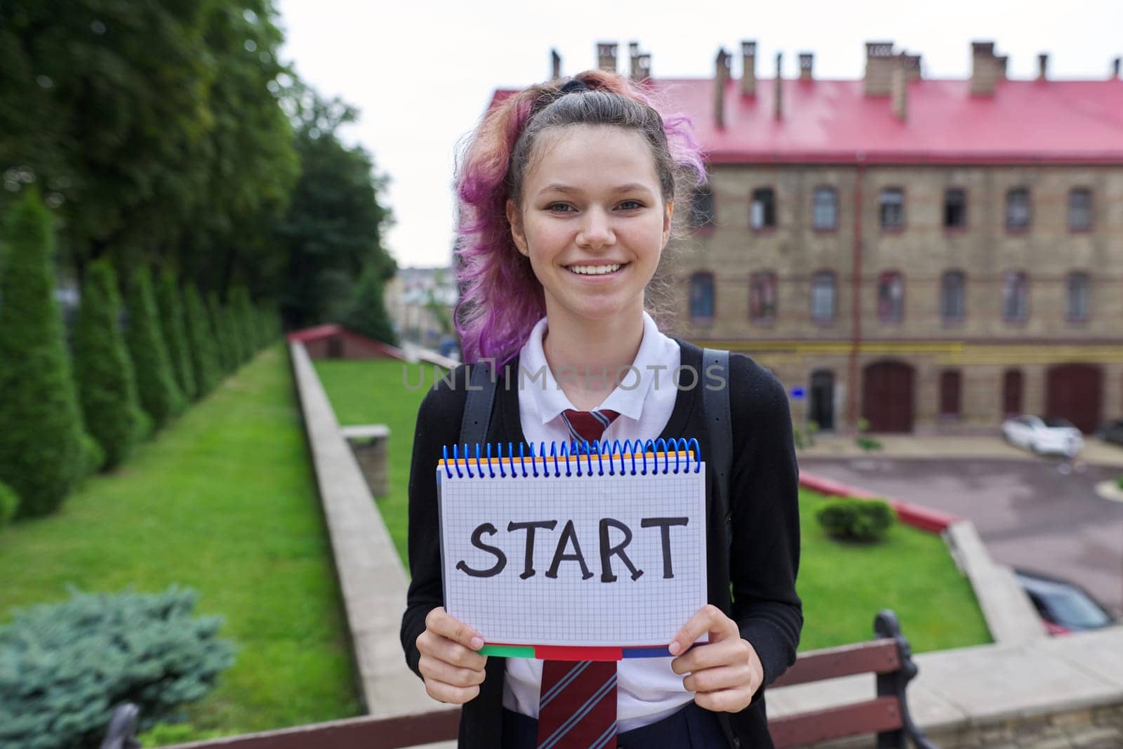 Teenager girl in uniform holding notepad with word start. Back to school, back to college, beginning of school year. Schoolgirl looking at camera, school building background