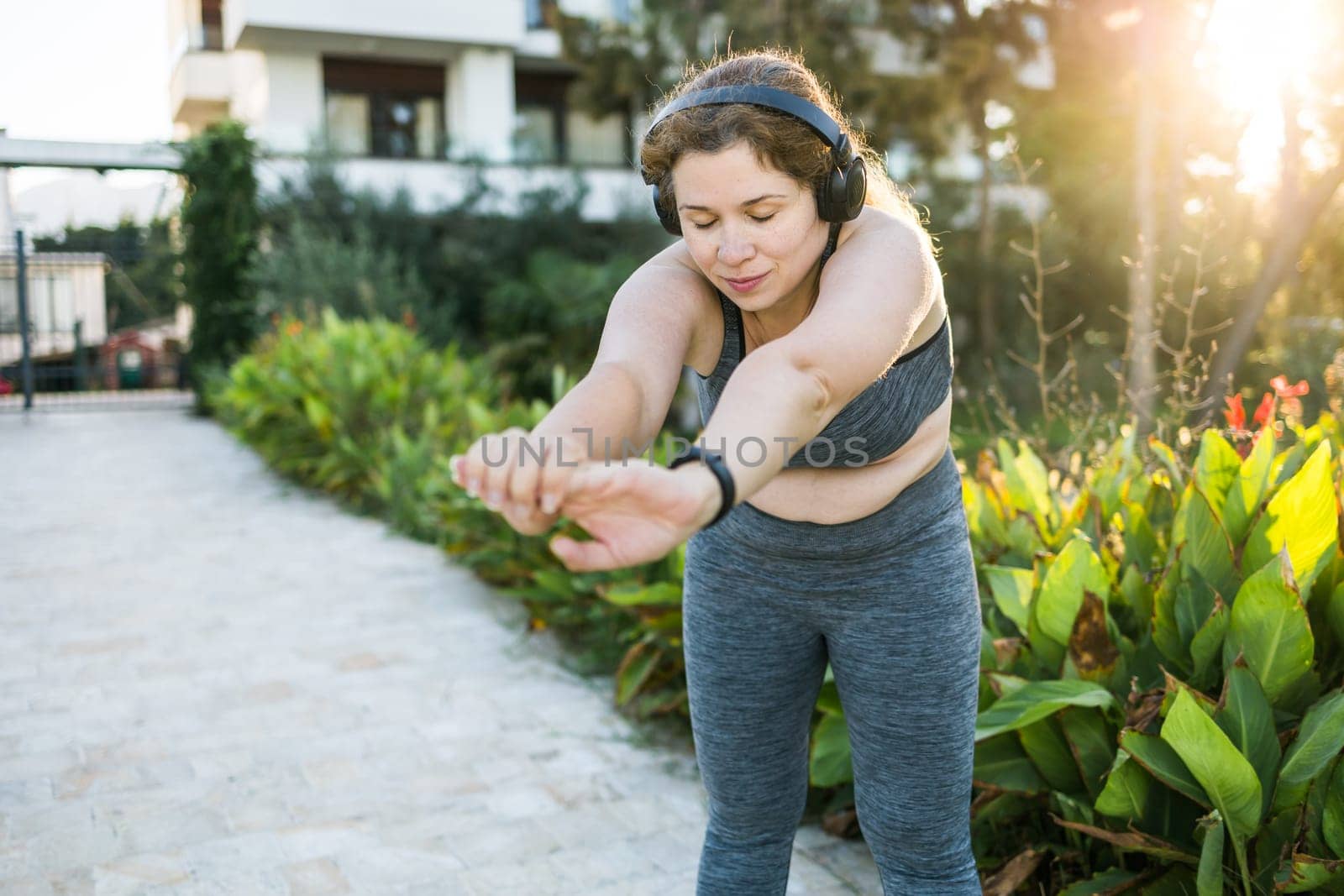 Adorable fat woman in tracksuit is engaged in fitness outdoor side view portrait. Young overweight woman lunges outdoors on warm summer day. Healthy lifestyle and weight loss.