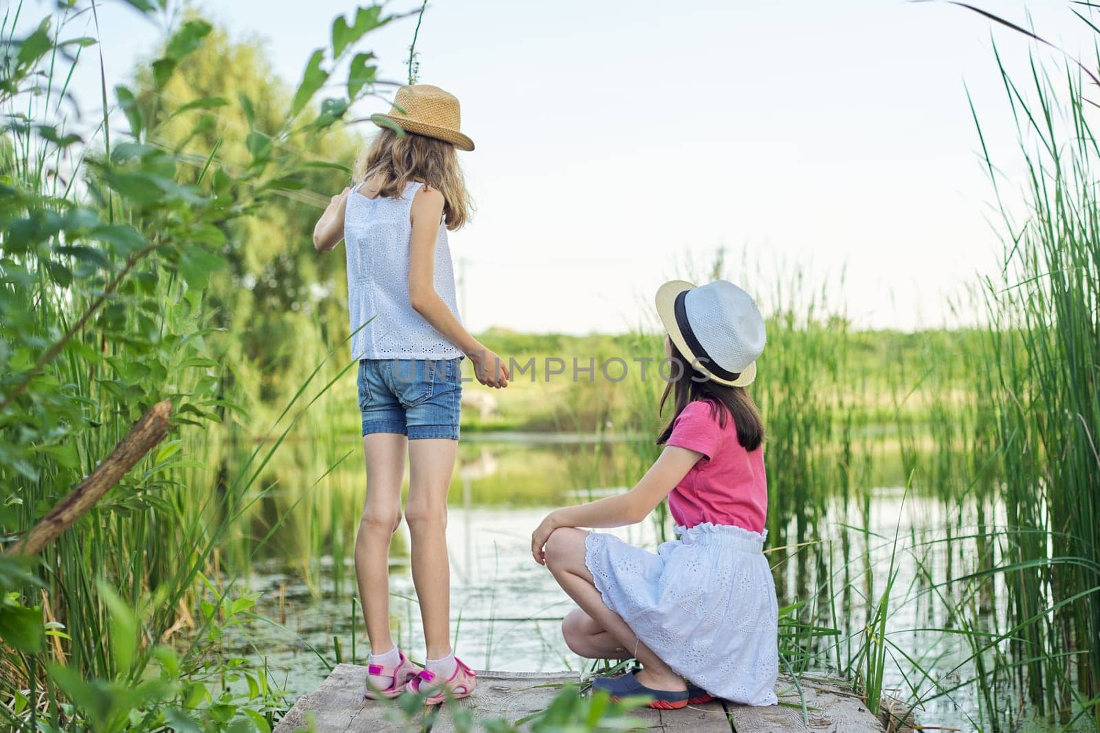 Two pretty girls children sitting on wooden pier of the lake in reeds, playing with water, talking, back view. Summer holidays, nature, happy childhood, friendship, country style.