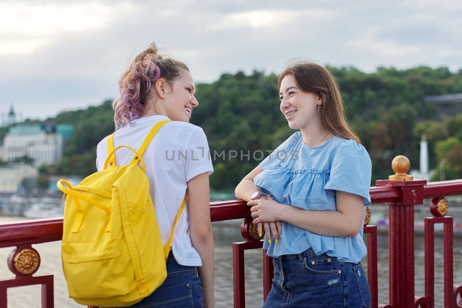 Portrait of two teenage girls standing on bridge over river, friends walk on summer sunny day. Friendship, lifestyle, youth, teens