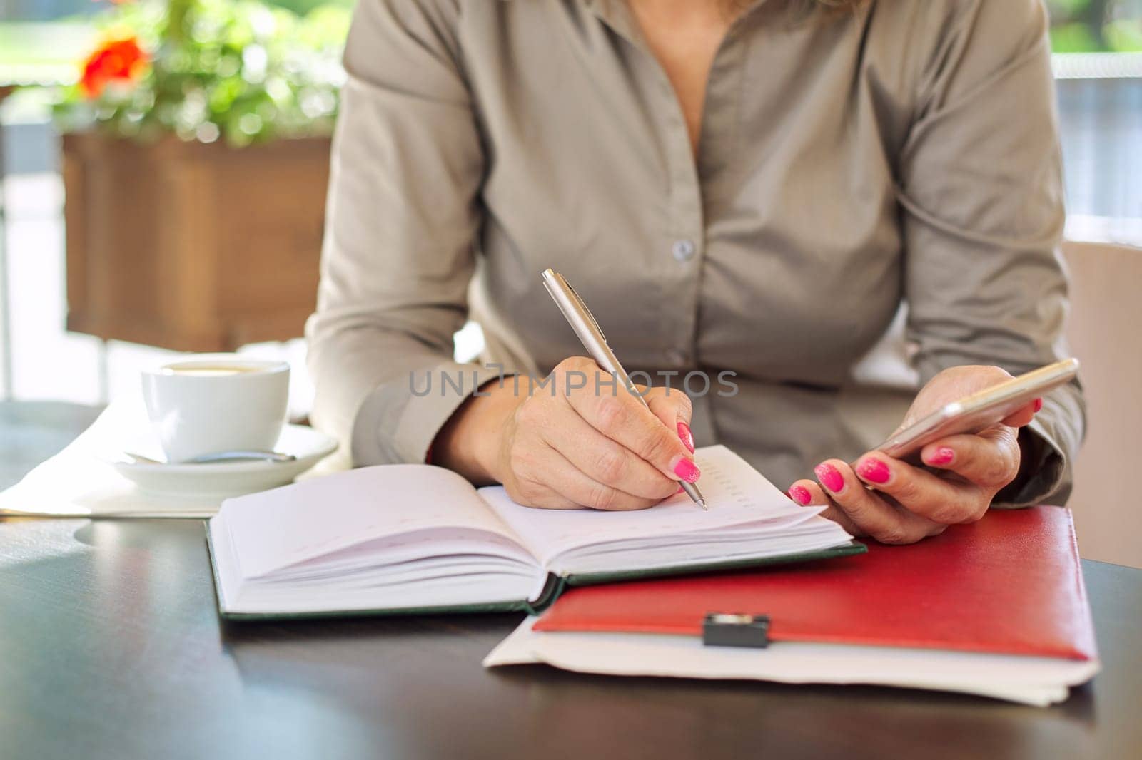Businesswoman writing in business notebook using smartphone, close up, female sitting in summer outdoor cafe, coffee break