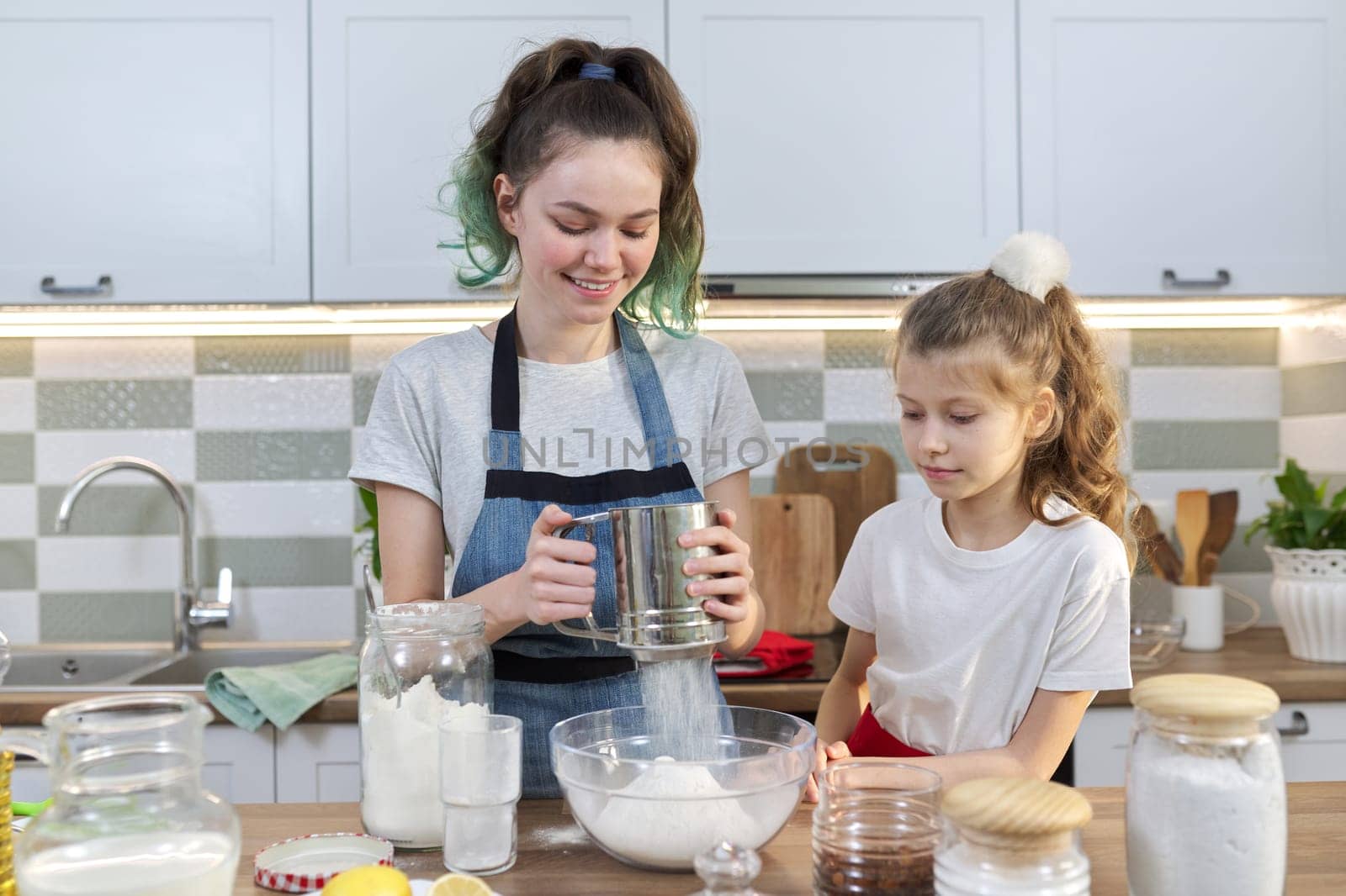 Two girls, teenager and younger sister, preparing cookies together in kitchen. Kids stir flour, the eldest shows the youngest. Family, friendship, fun, healthy homemade food