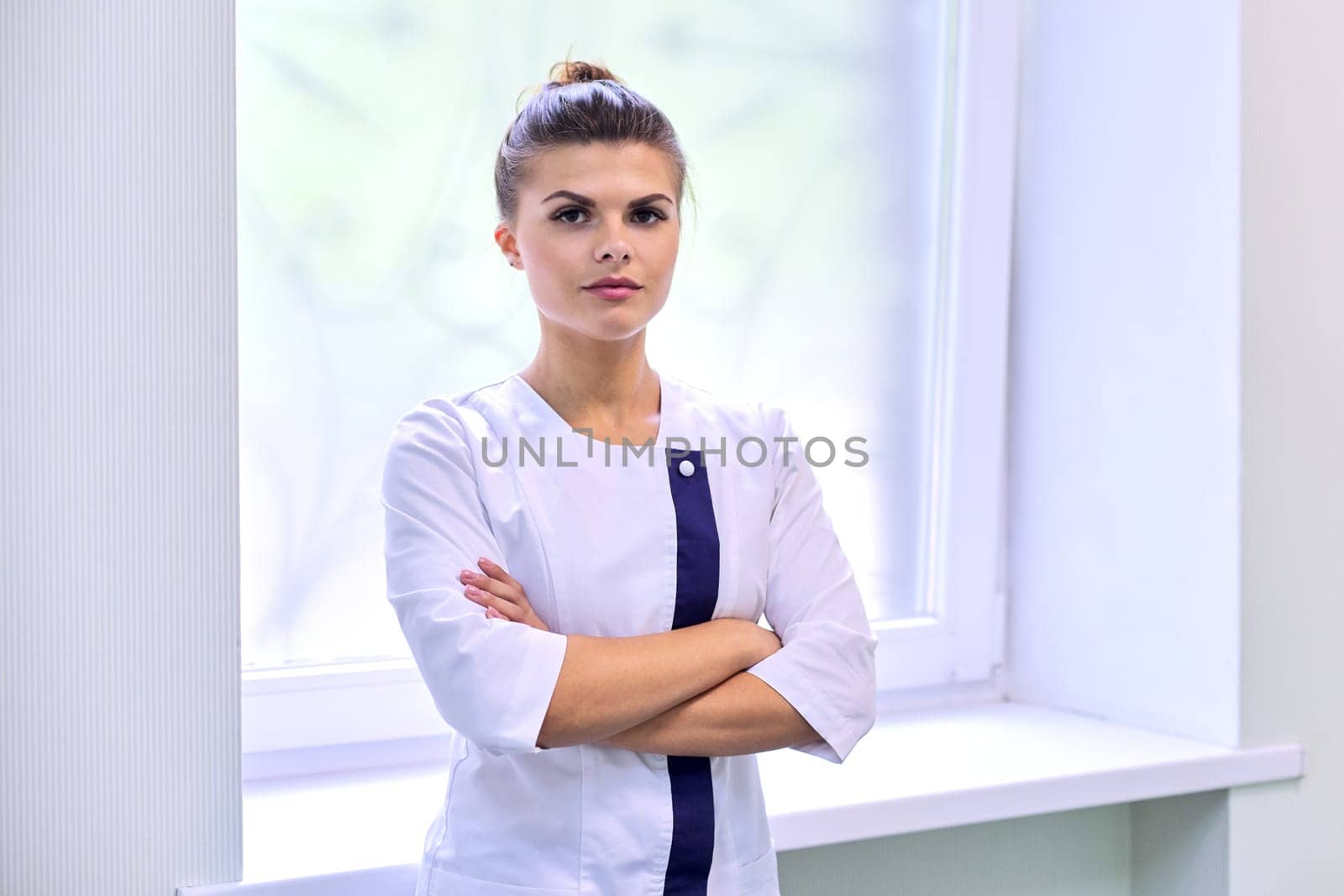 Portrait of young attractive confident woman in medical uniform with arms crossed, light wall clinic window background. Female pharmacist, nurse, cosmetologist, specialist, scientist