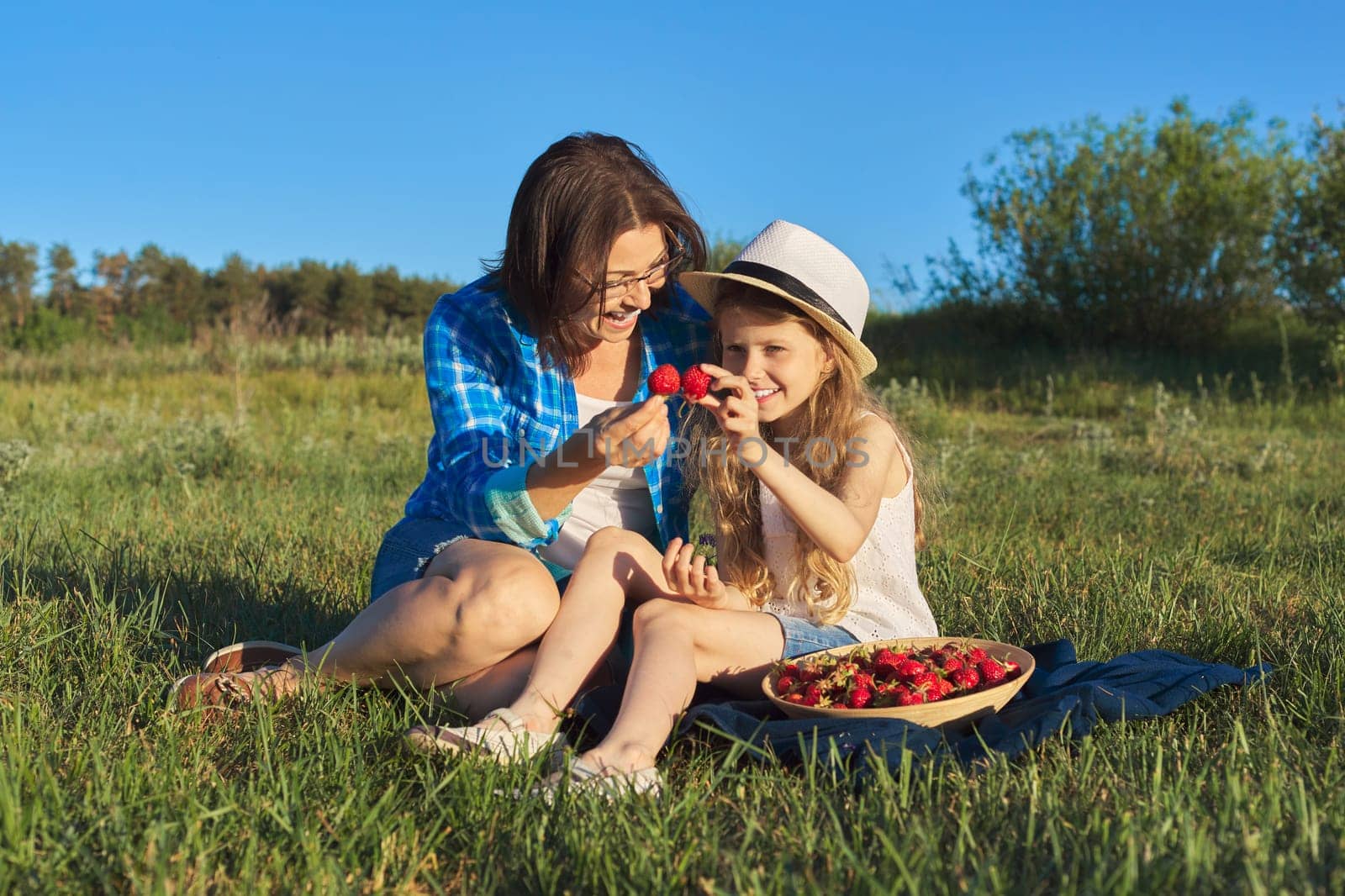 Happy laughing mother and child daughter together outdoors, sitting on green grass, picnic with bowl of fresh strawberries. Mothers day, May nature