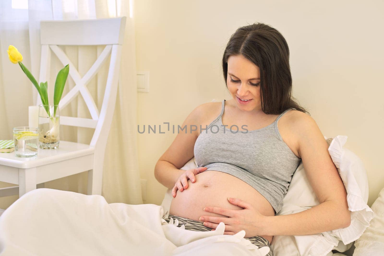 Young beautiful pregnant woman sitting at home in bed, female touching her tummy, waiting for the birth of baby