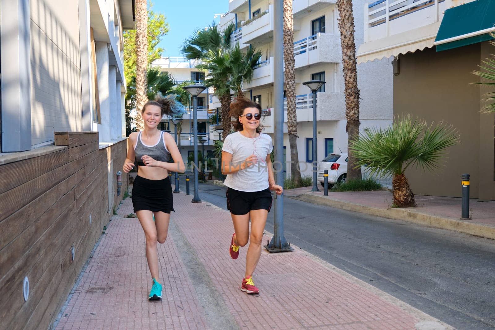Active friendly sportive family, mother and daughter teenager running together. Tropical city street background, beautiful happy jogging women
