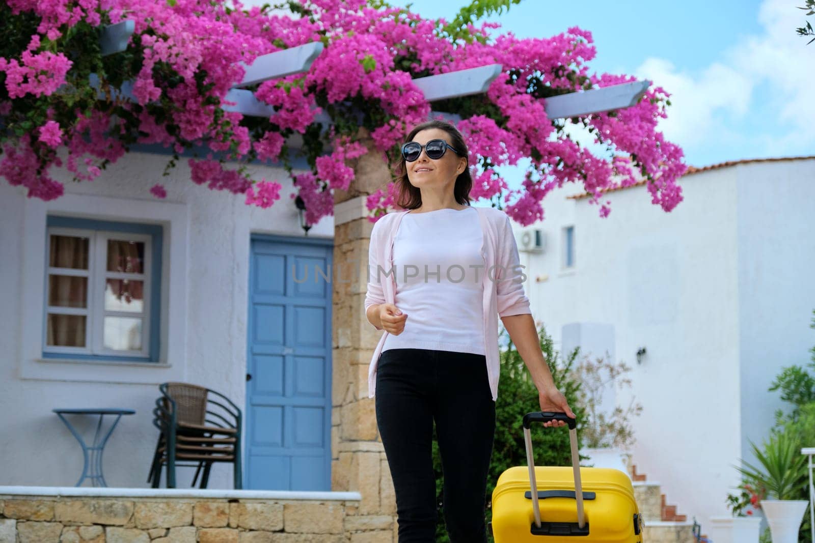 Woman with suitcase walking outdoor through territory of sea picturesque resort spa hotel with beautiful pink flower landscaping. Travelling, vacation, leisure, weekend, people
