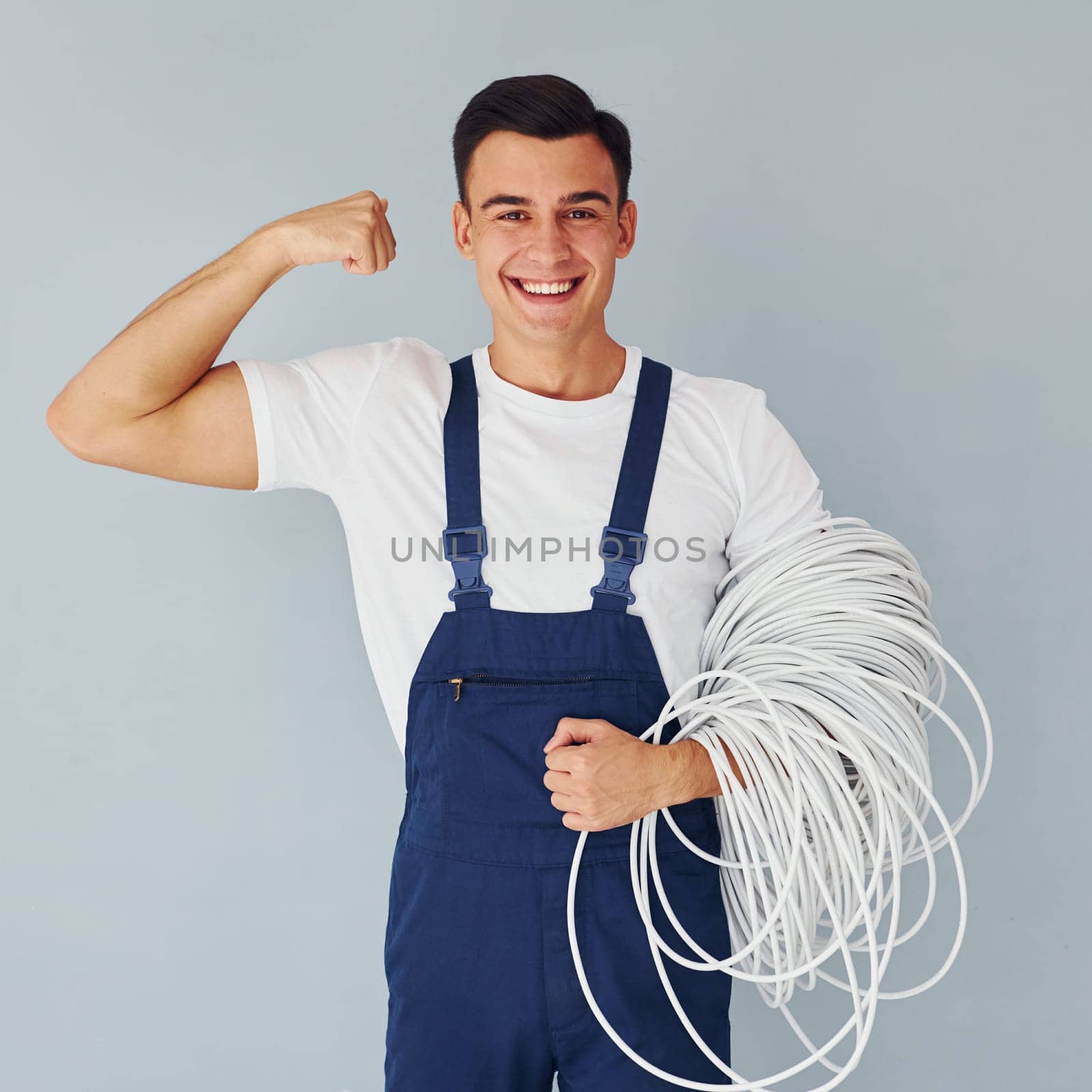Holds cable. Male worker in blue uniform standing inside of studio against white background.