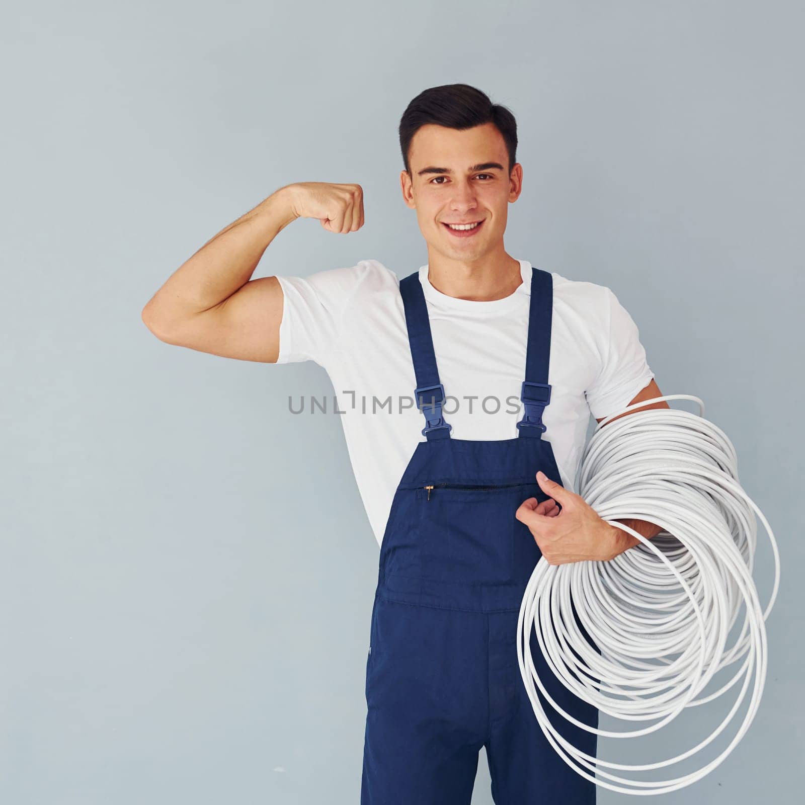 Holds cable. Male worker in blue uniform standing inside of studio against white background.