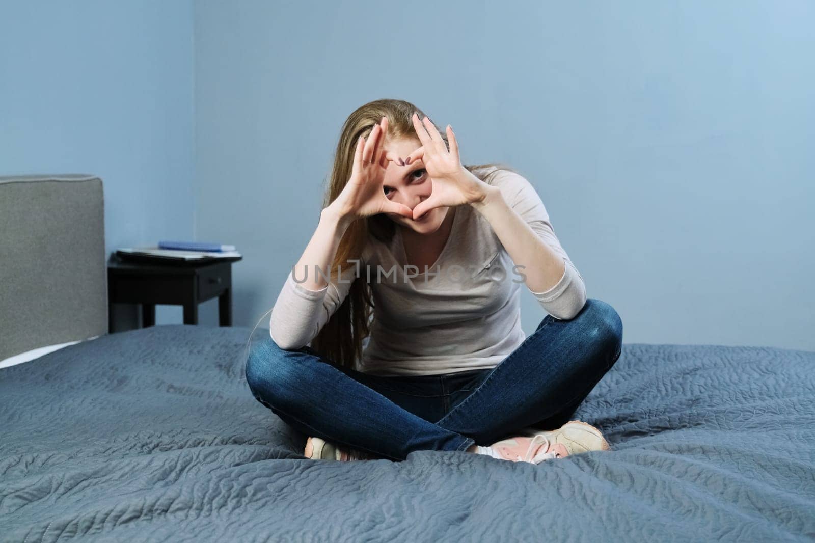 Positive beautiful young woman showing hands sign symbol heart, sitting at home on the bed. Emotion love, happiness, joy, valentines day, young people concept