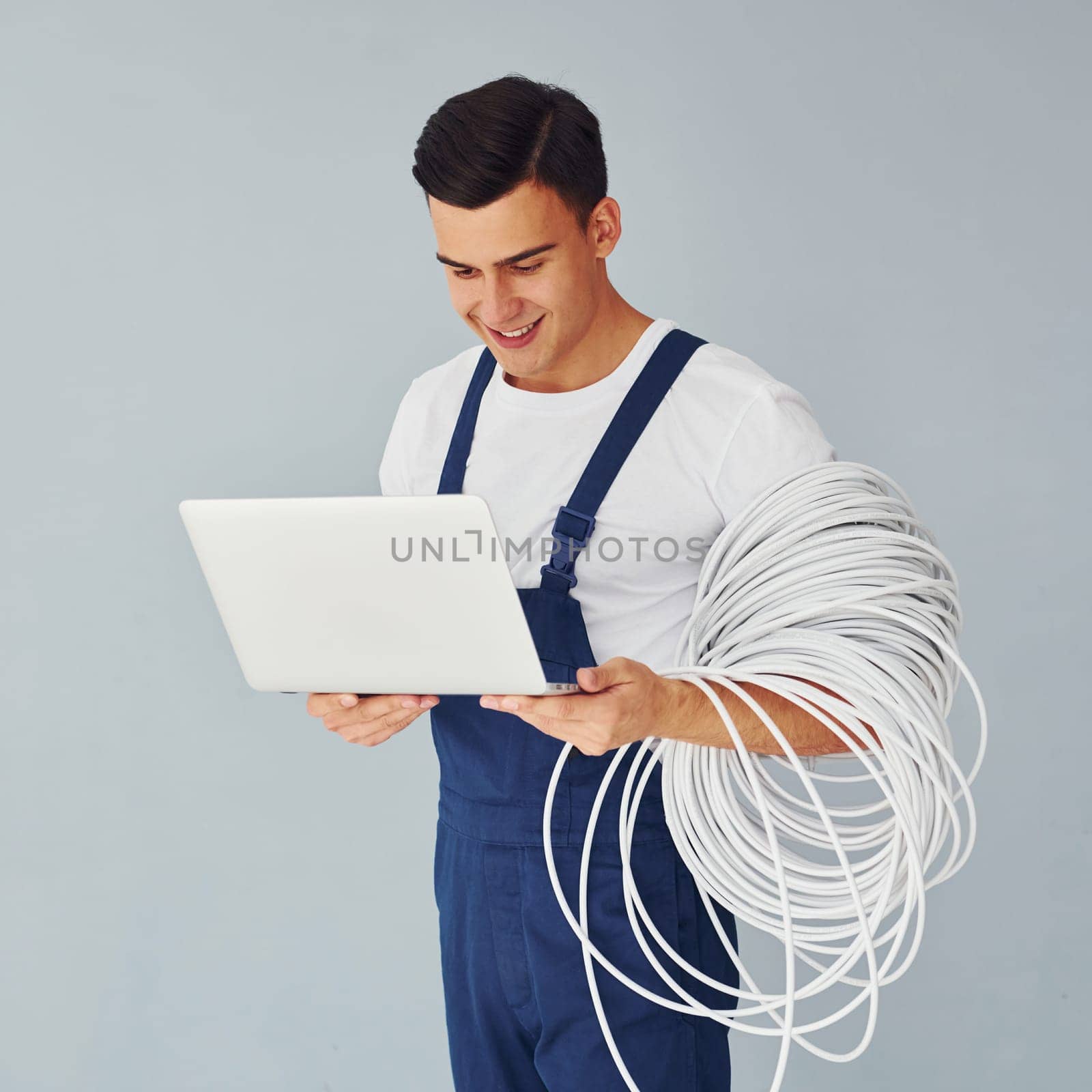 Uses laptop. Male worker in blue uniform standing inside of studio against white background.