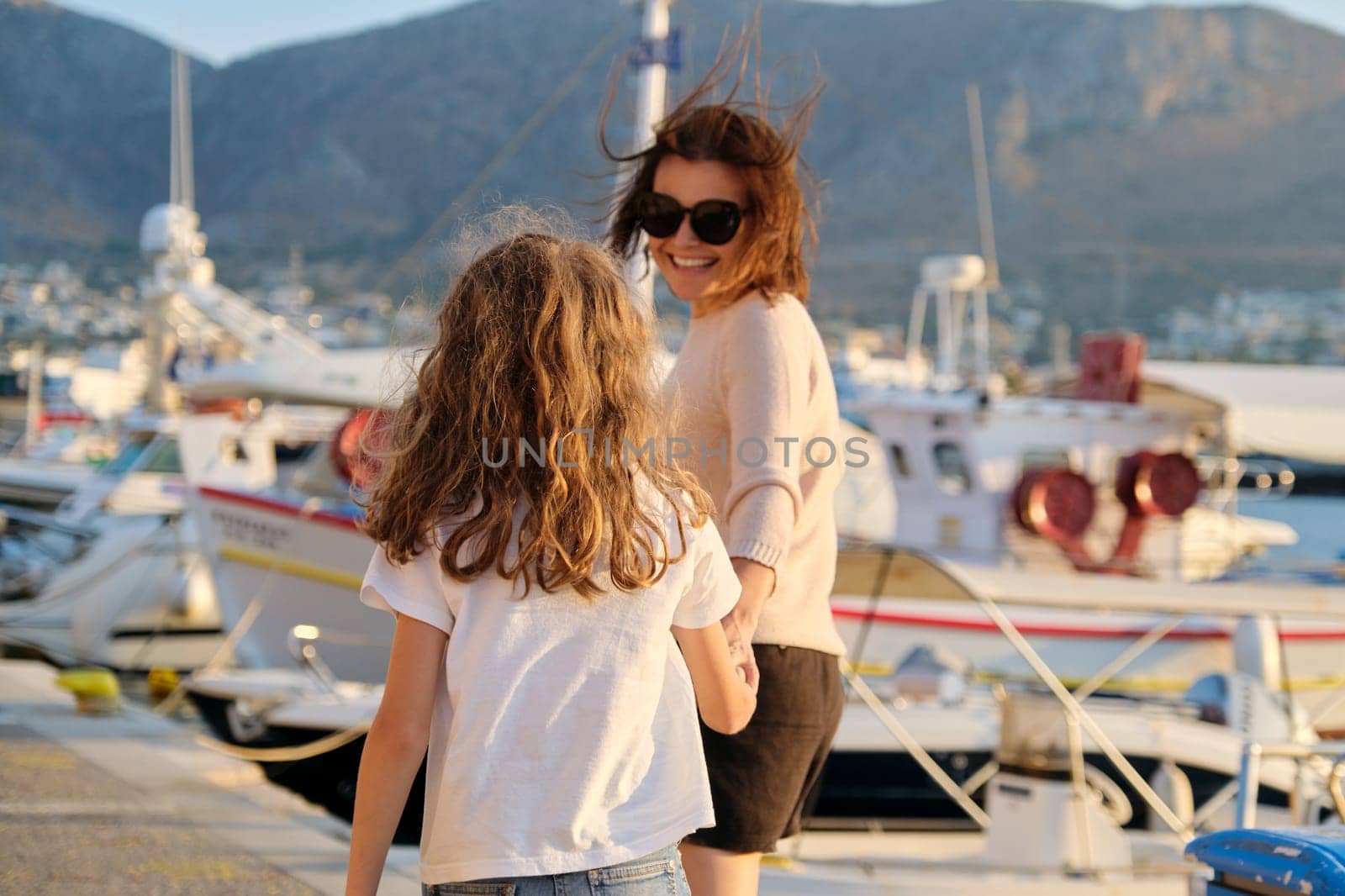 Happy mother and daughter child walking together holding hands along seafront. Marina for yachts, sunset on sea, scenic mountains background. Family, love, travel, lifestyle, vacation concept