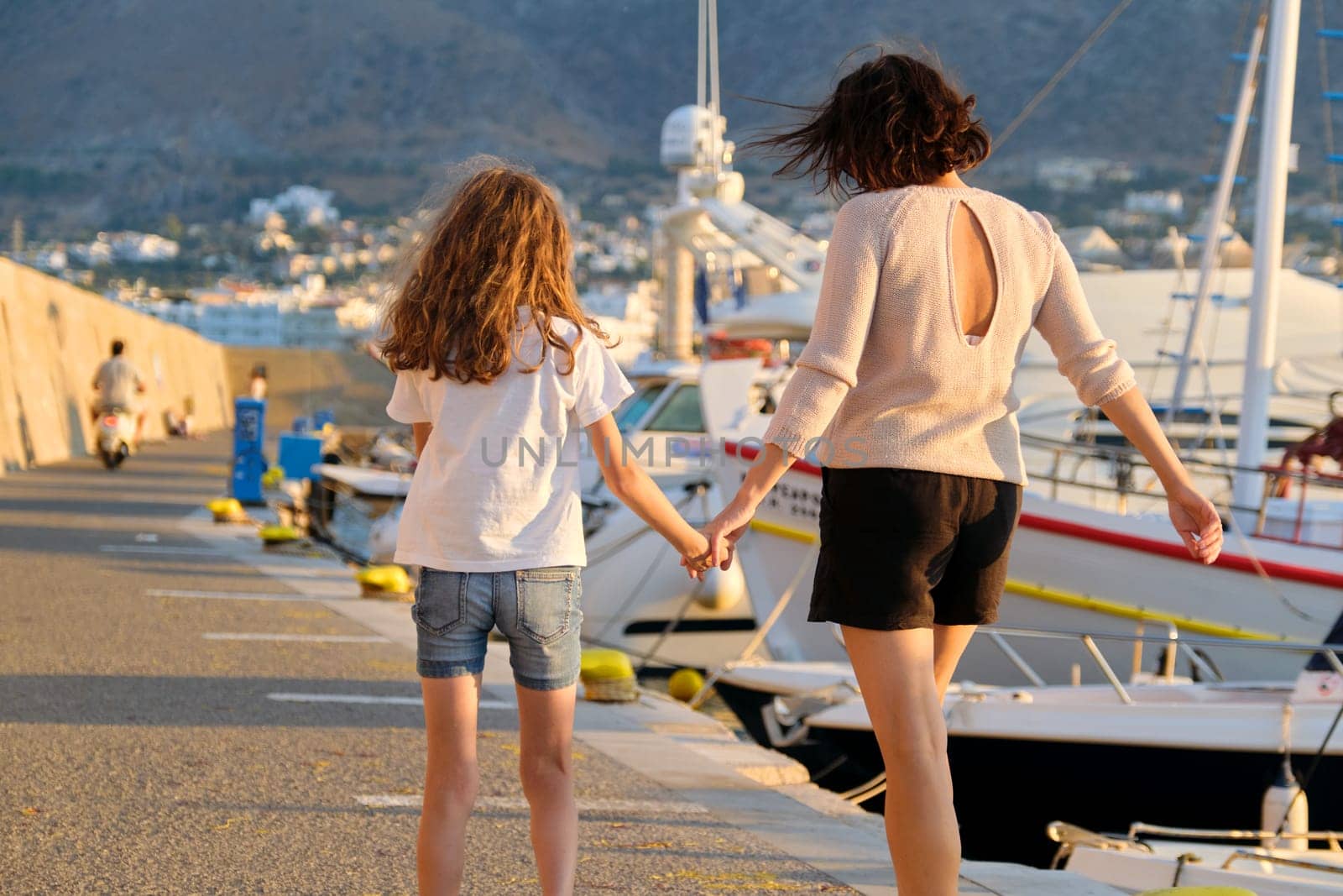 Happy mother and daughter child walking together holding hands along seafront. Marina for yachts, sunset on sea, scenic mountains background. Family, love, travel, lifestyle, vacation concept