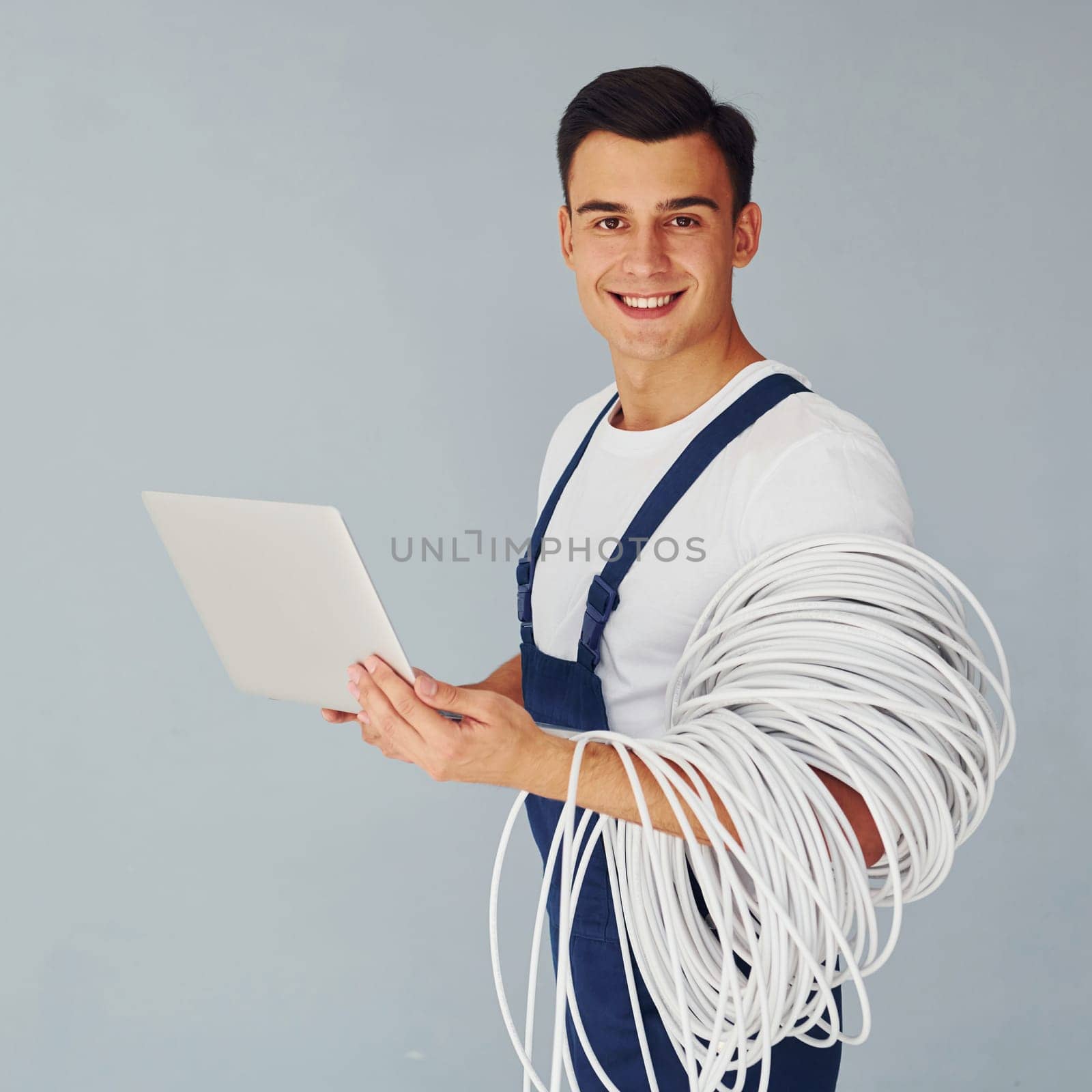 Uses laptop. Male worker in blue uniform standing inside of studio against white background.