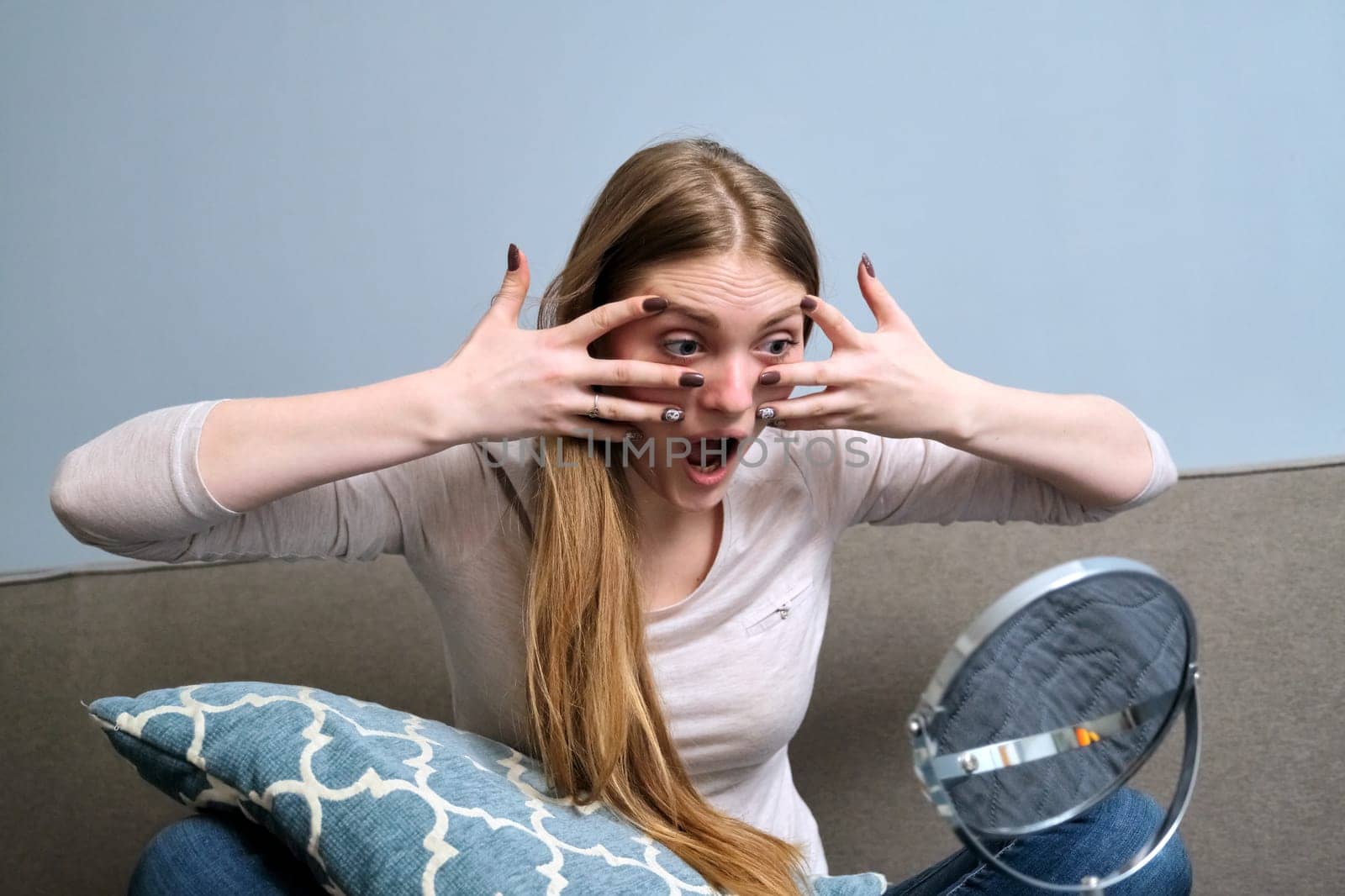 Young beautiful woman with make-up mirror doing face massage with hands. Girl sitting at home in bed. Beauty, youth, skin care