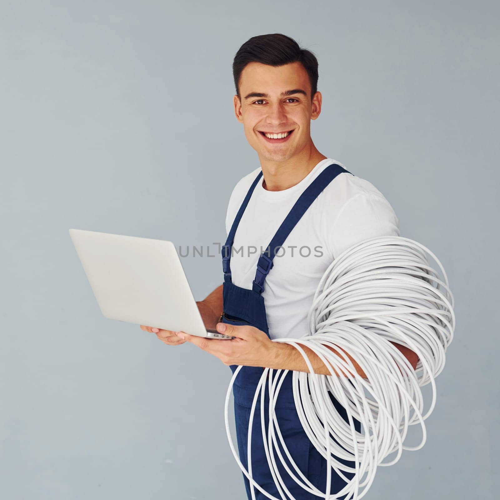 Uses laptop. Male worker in blue uniform standing inside of studio against white background.