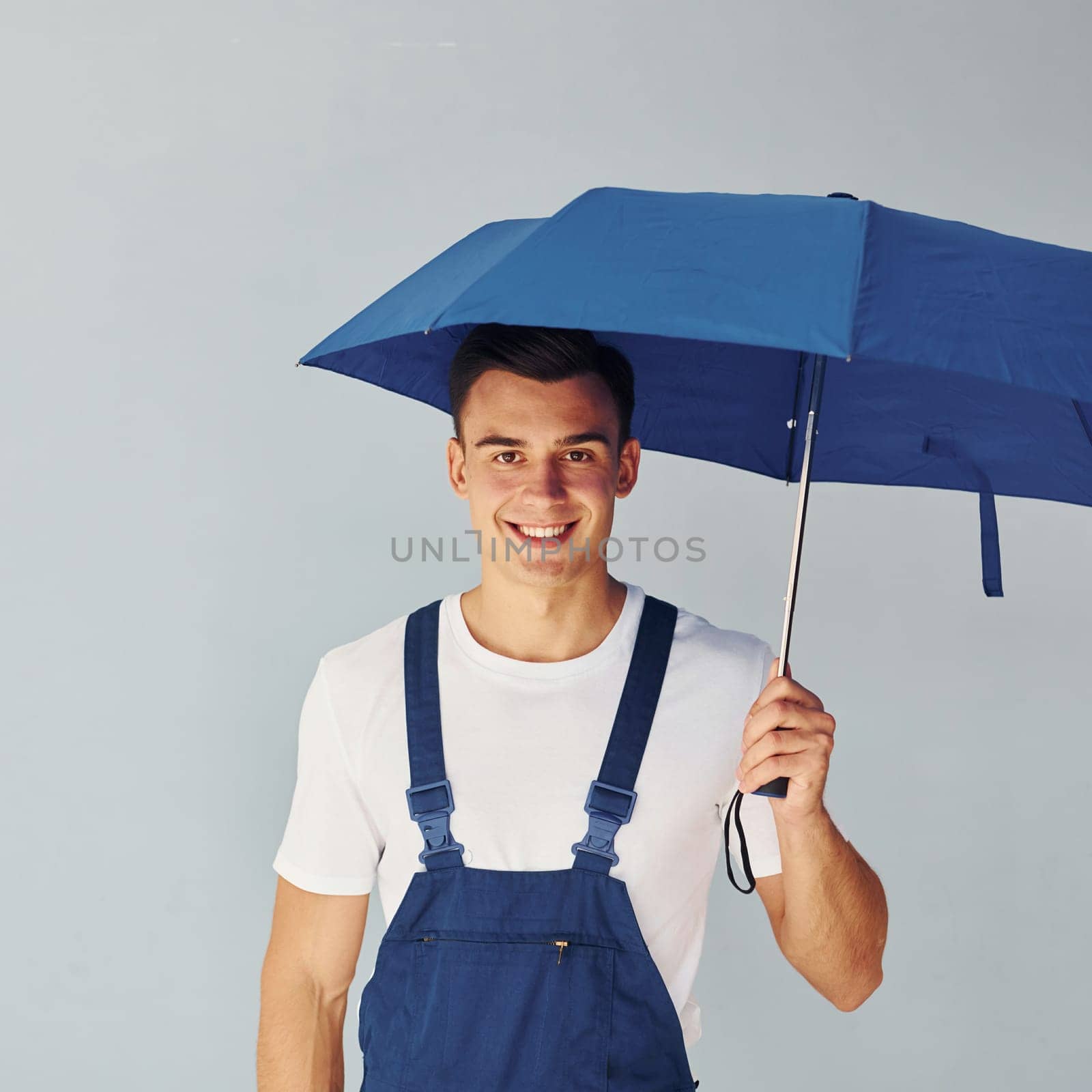 Holds umbrella by hand. Male worker in blue uniform standing inside of studio against white background.