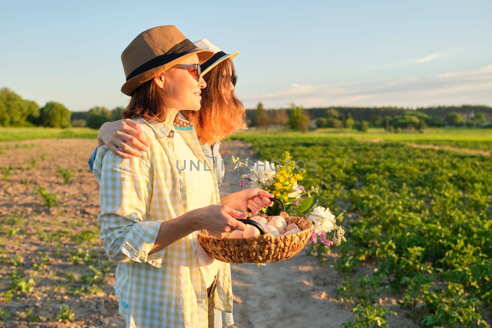 Portrait of two happy smiling embraced women mother and teenage daughter with basket of farm eggs in hats, with bouquet of flowers in garden, summer nature, sunset background, copy space