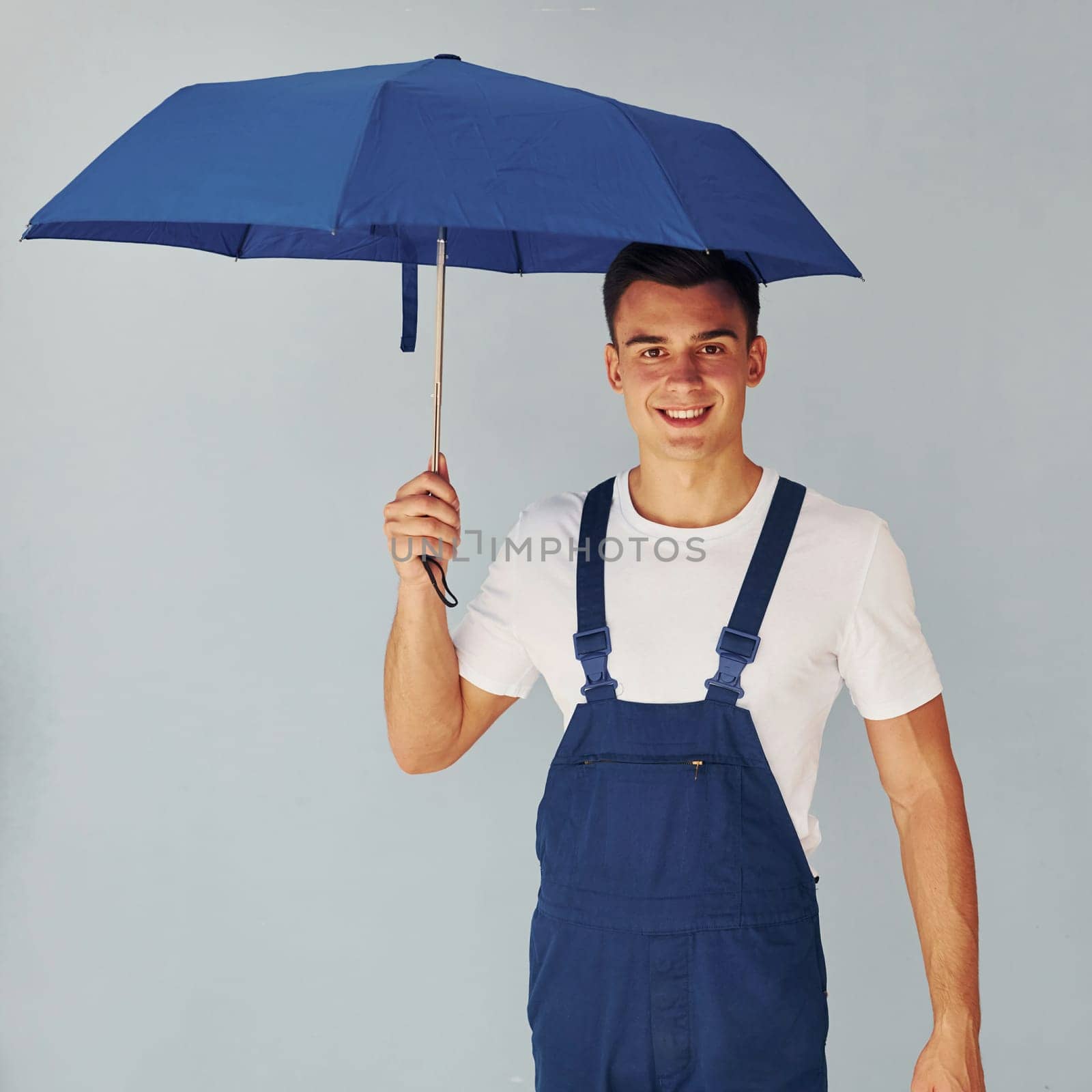 Holds umbrella by hand. Male worker in blue uniform standing inside of studio against white background.